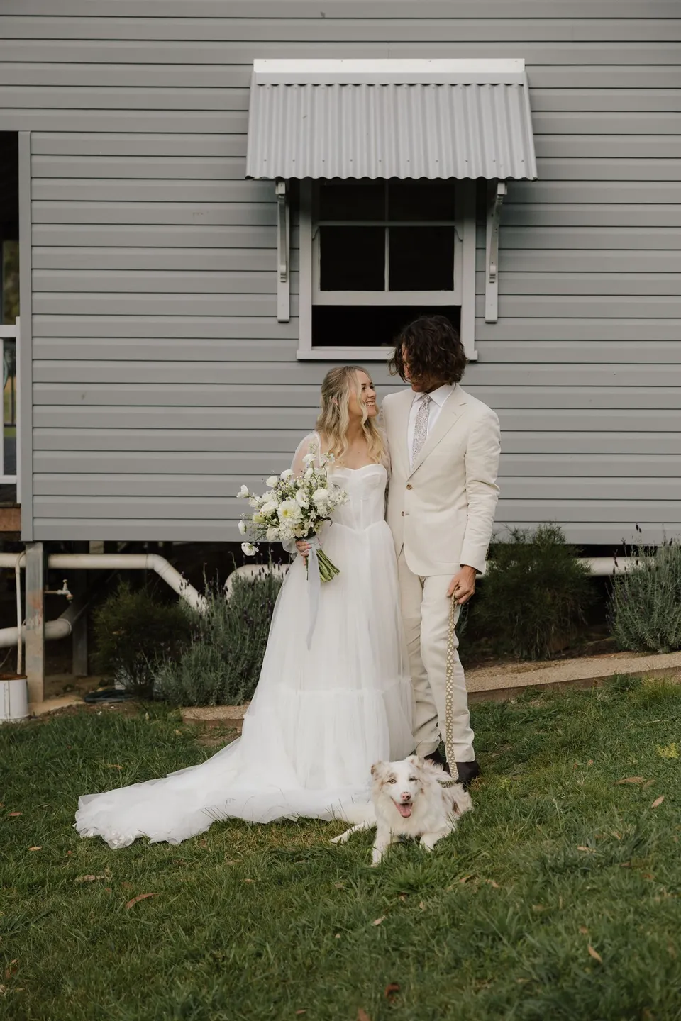 Bridget and James standing beside the weatherboard cottage with a small white dog at their feet. At Cudgera Creek in northern New South Wales, for an Elopement Collective elopement. Photograph by Bulb Creative.