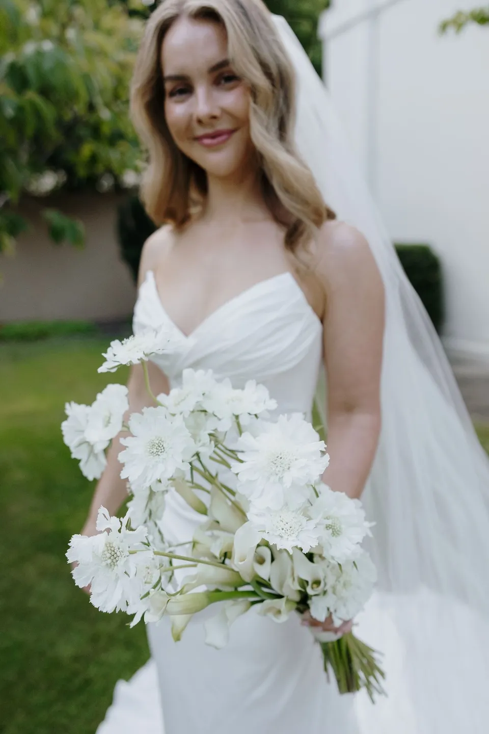 Tanesha smiling in the garden with her white bouquet by The Romantics held in front of her wedding dress, at Islington Hotel in South Hobart, Tasmania, Australia, for an Elopement Collective summer elopement. Photograph by George Bowden.