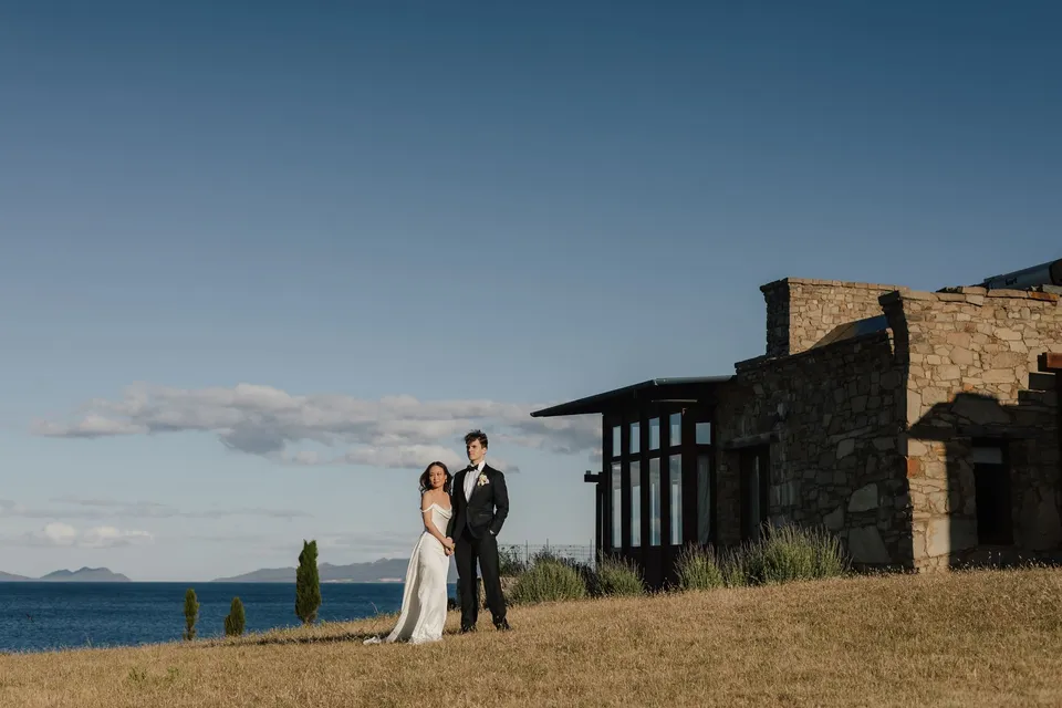 Clara and Harry holding hands beside Thalia Haven’s stone house, with golden grassland, cypress trees and blue water around them, at Thalia Haven on the East Coast of Tasmania, for an Elopement Collective elopement. Photograph by Bulb Creative.