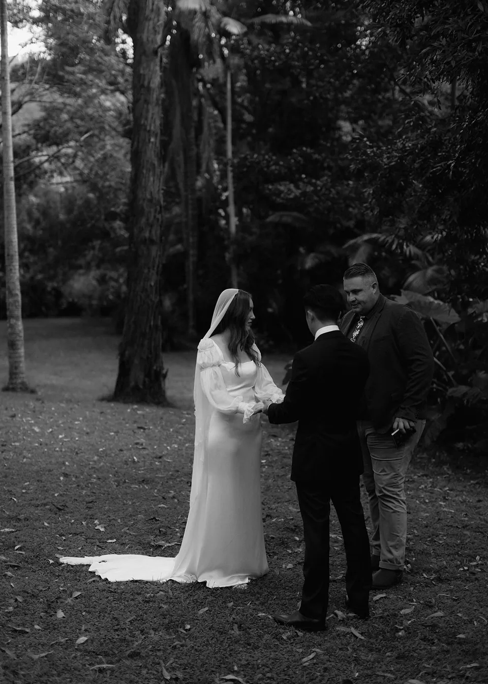 Wide black-and-white view of the couple and celebrant Josh Withers on the lawn. At Fig Tree Villa in Ewingsdale, Byron Bay, for an Elopement Collective elopement. Photograph by Bulb Creative.