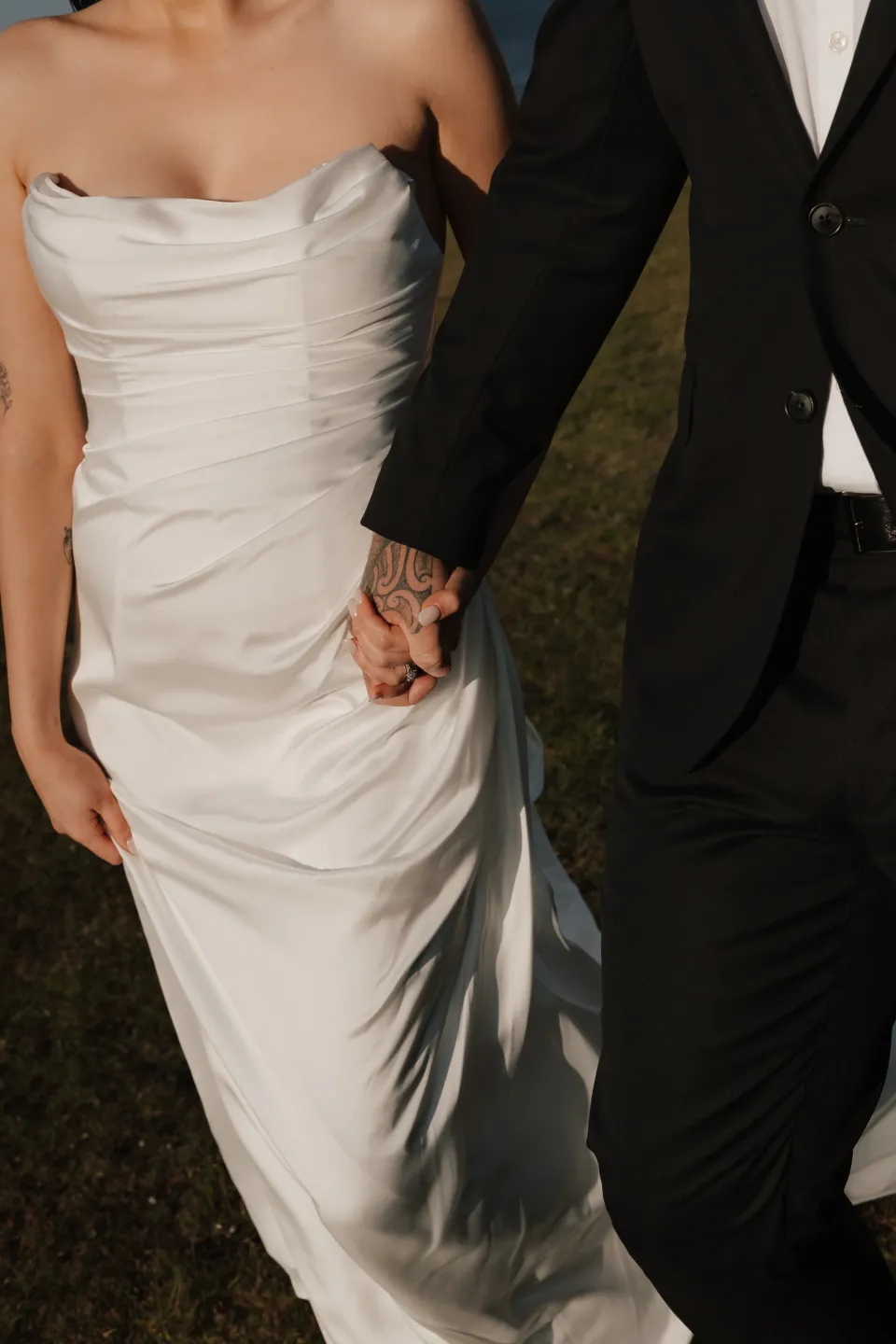 Close view of Dani and Anthony holding hands, showing her draped white dress and his dark suit, at Thalia Haven on Tasmania’s east coast, for an Elopement Collective elopement. Photograph by Bulb Creative.