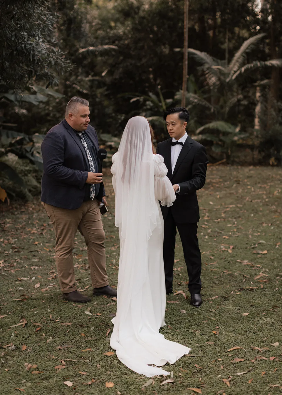 Elyse and Jonathan facing each other with celebrant Josh Withers beside them. At Fig Tree Villa in Ewingsdale, Byron Bay, for an Elopement Collective elopement. Photograph by Bulb Creative.