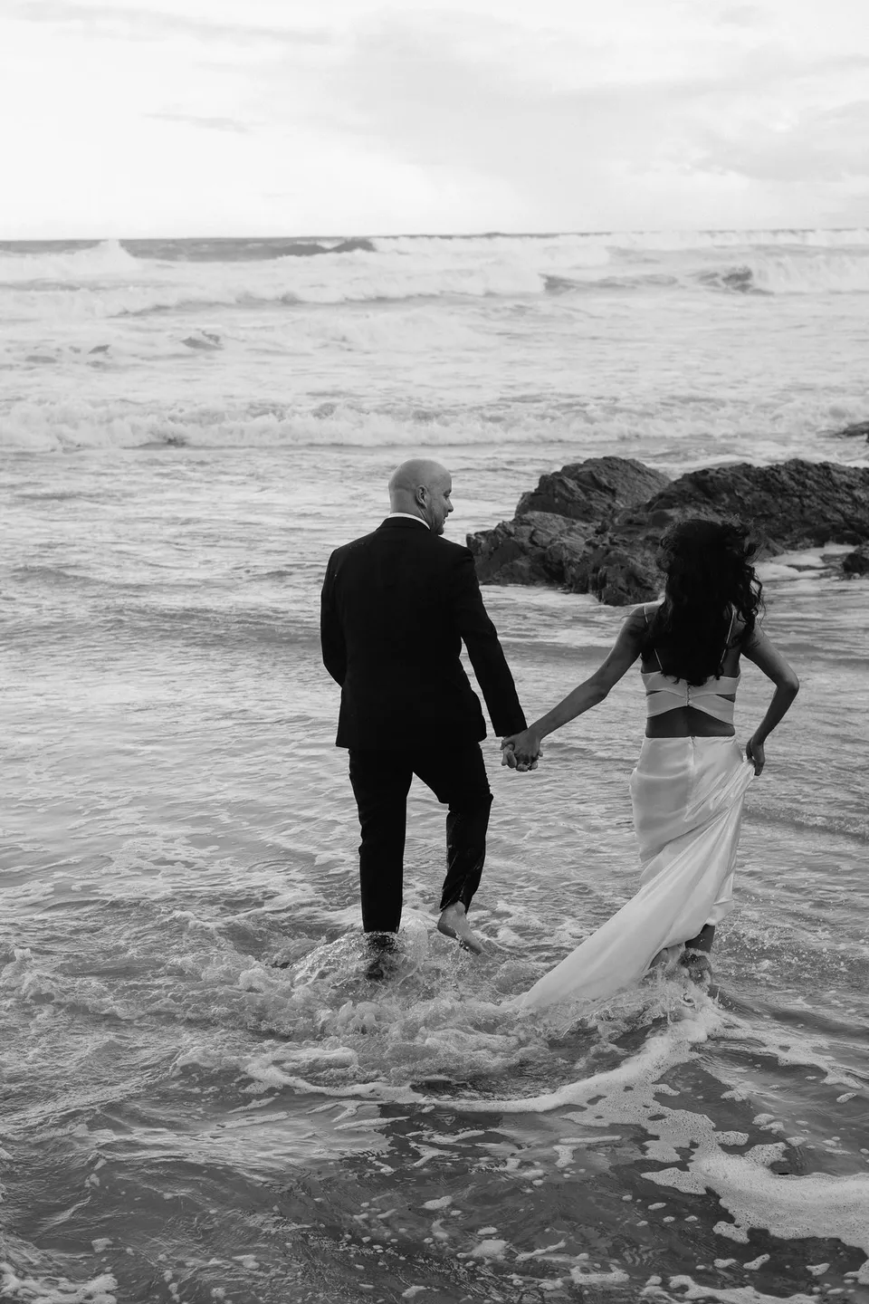 Black-and-white photograph of Rachel and Dane stepping into the wash, her dress trailing behind her. At Whites Beach, Broken Head, near Byron Bay, for an Elopement Collective elopement. Photograph by Jason Lucas.