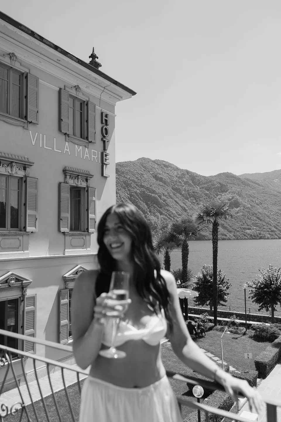 Black and white photograph of Lyndal holding a glass on the balcony at Hotel Villa Marie in Tremezzo, with the hotel facade, lake and mountains behind her, at Lake Como in Lombardy, Italy, for an Elopement Collective elopement. Photograph by George Bowden.