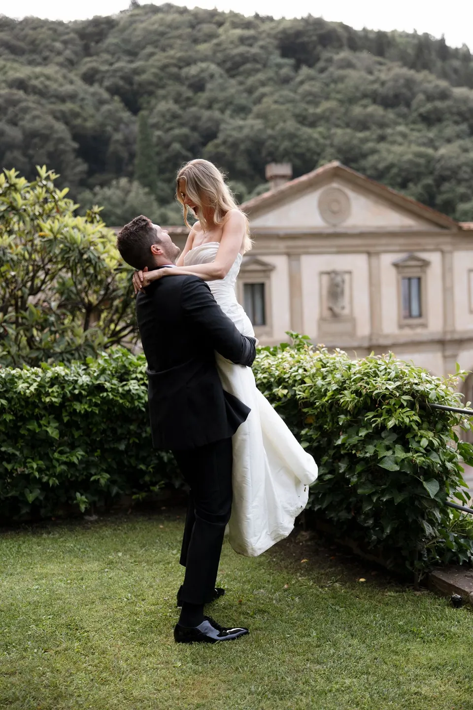 Matthew lifting Holly into his arms on the garden lawn with the villa facade behind them, at Villa San Michele in Fiesole above Florence, Tuscany, Italy, for an Elopement Collective elopement. Digital photograph by Khara from House of Lucie.