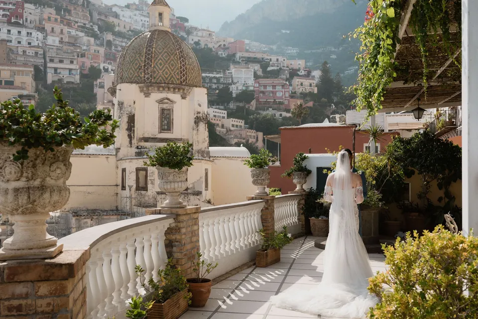 Skyler’s veil and lace dress from behind as she faces Zach on the terrace, beside flower-filled urns and the tiled church dome, at Hotel Buca di Bacco in Positano, Amalfi Coast, Italy, for an Elopement Collective elopement. Photograph by Jason Corroto.
