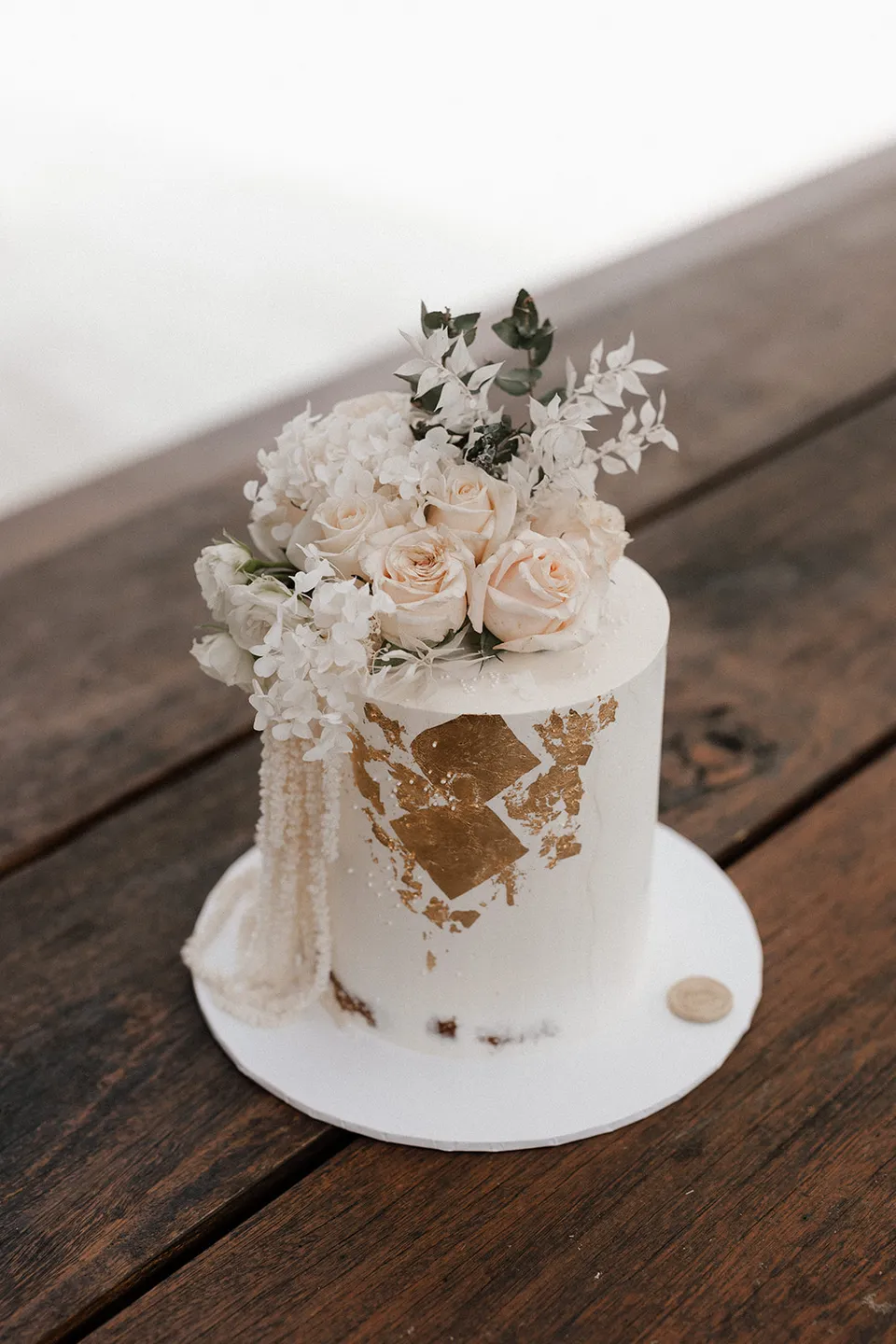 A small white wedding cake with gold detailing and pale flowers on a timber table, at Grounds of Cabarita in Cabarita Beach, NSW, for an Elopement Collective elopement. Photo by Bulb Creative.