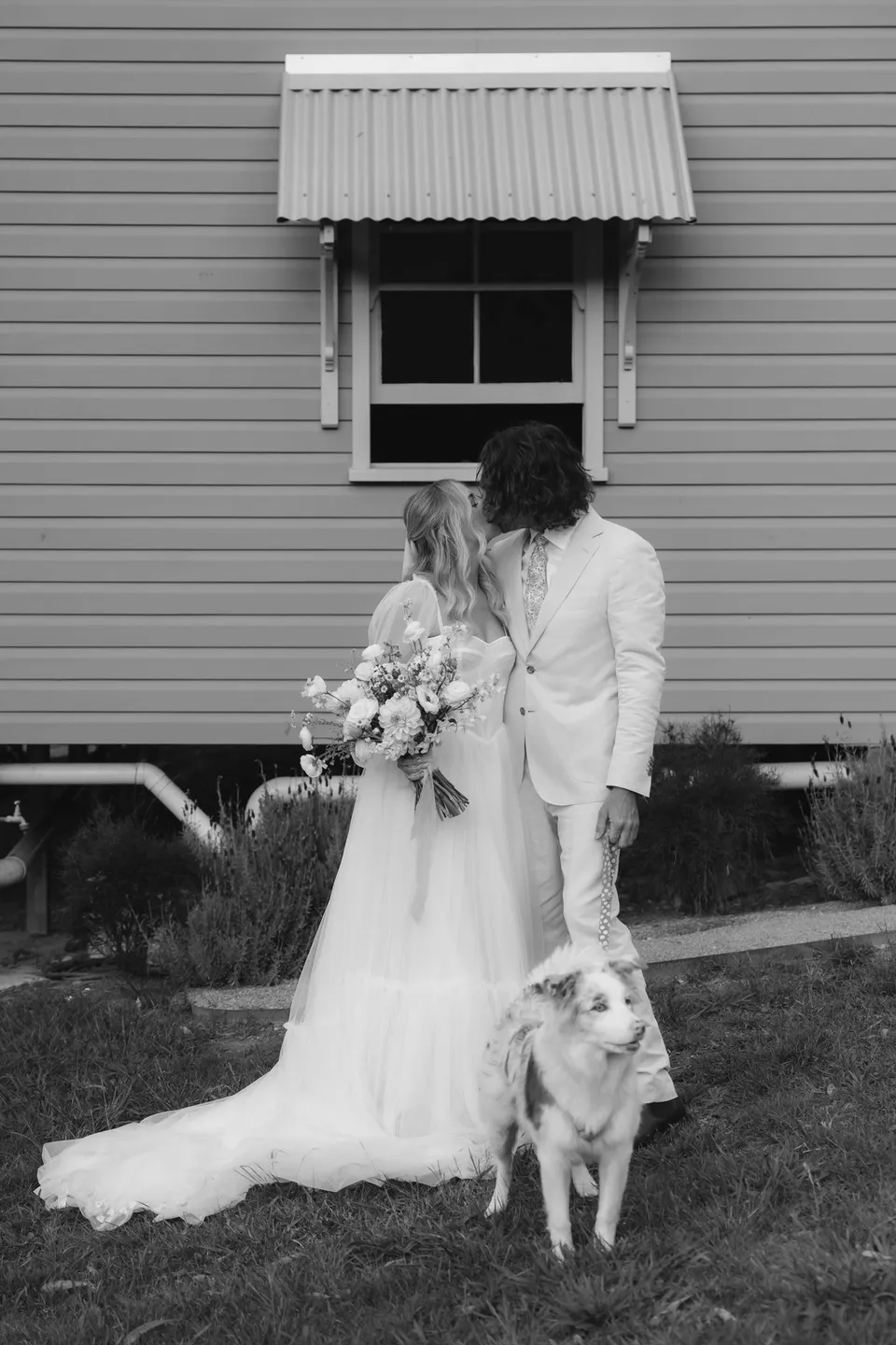 Black-and-white photograph of Bridget and James outside the weatherboard cottage with a small white dog. At Cudgera Creek in northern New South Wales, for an Elopement Collective elopement. Photograph by Bulb Creative.