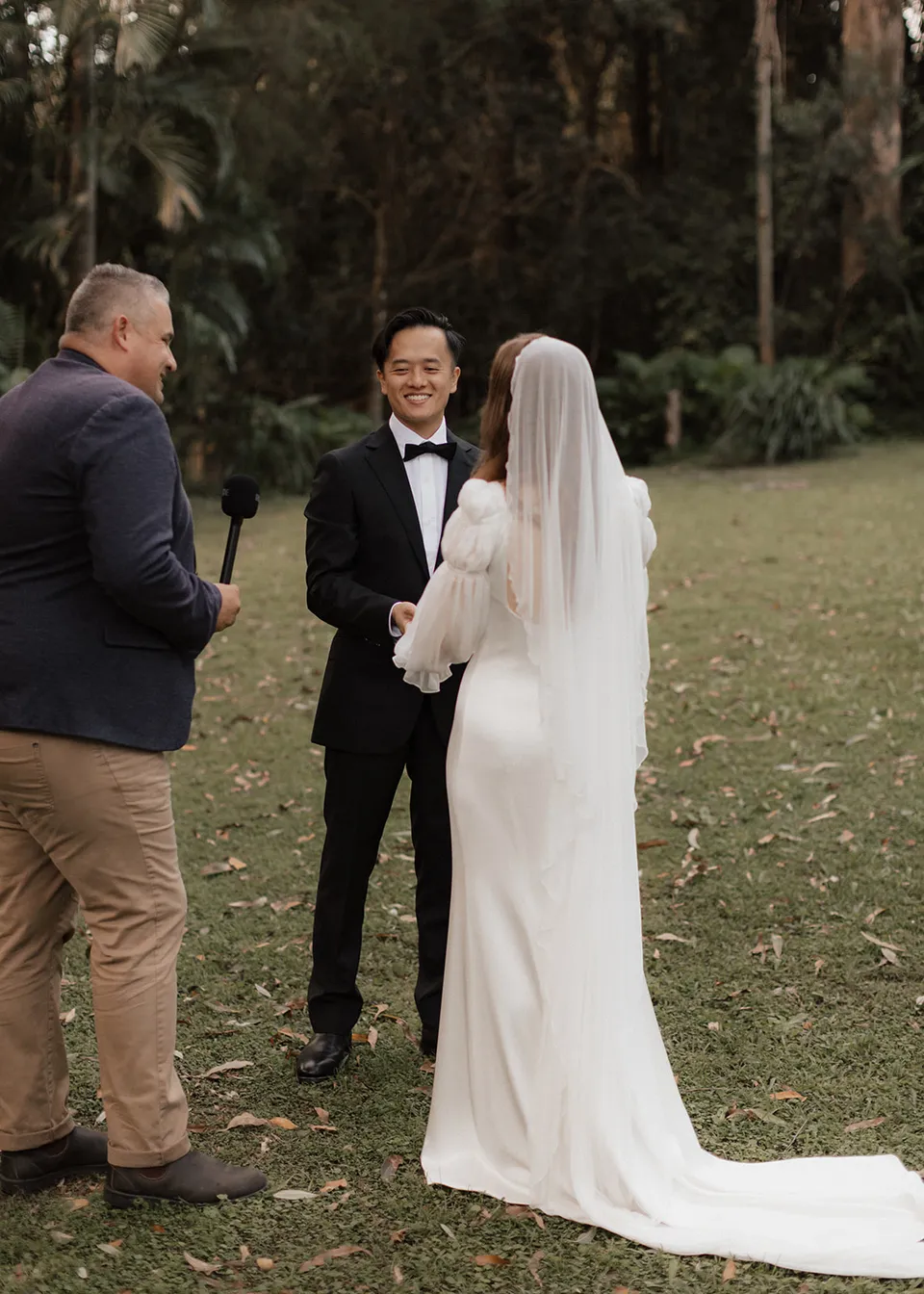Jonathan smiling at Elyse as celebrant Josh Withers speaks. At Fig Tree Villa in Ewingsdale, Byron Bay, for an Elopement Collective elopement. Photograph by Bulb Creative.