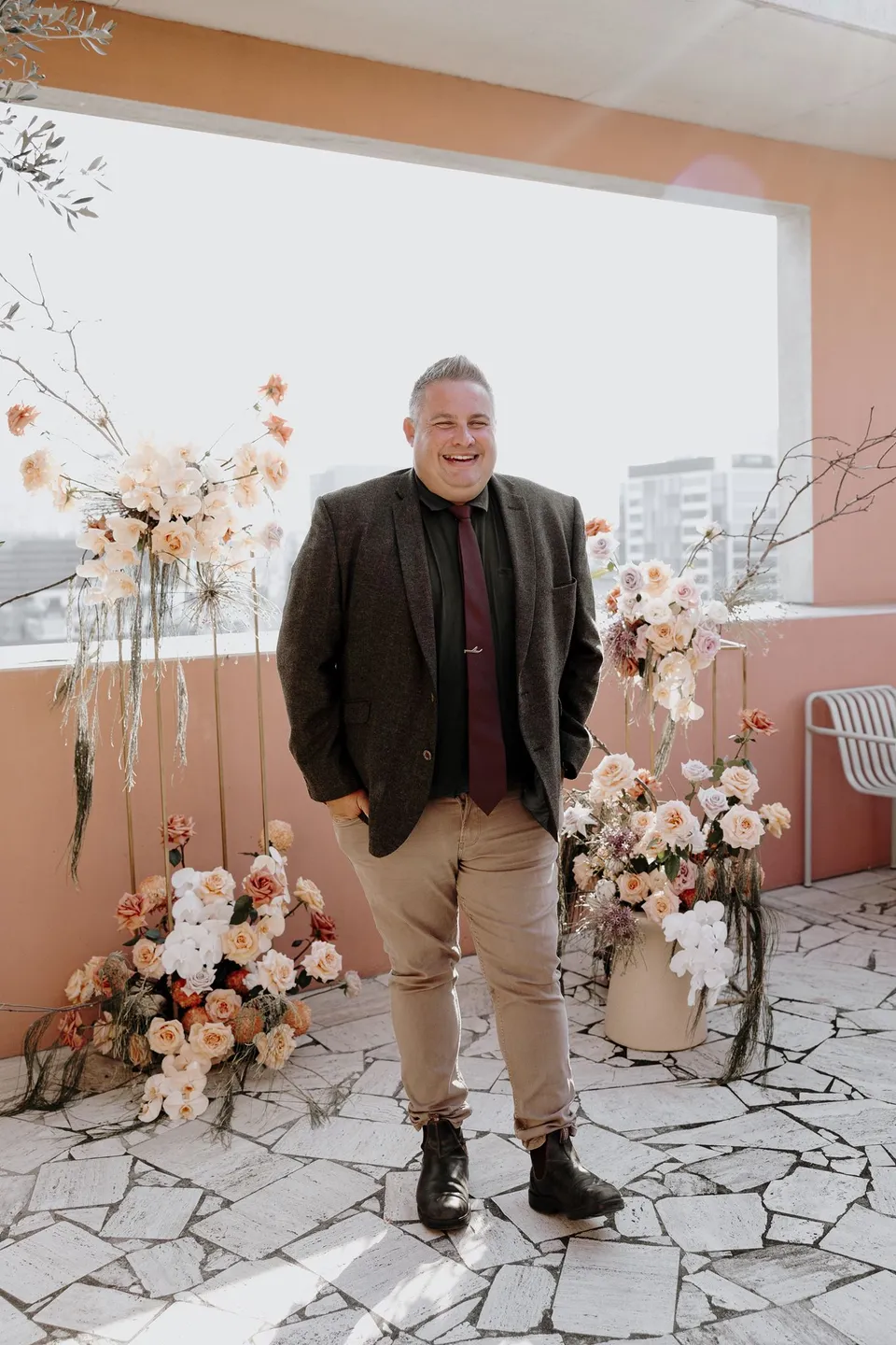 Celebrant Josh Withers standing between the terrace flower arrangements with Brisbane behind him, during Danielle and Adam’s Elopement Collective elopement at The Calile Hotel in Fortitude Valley, Brisbane.