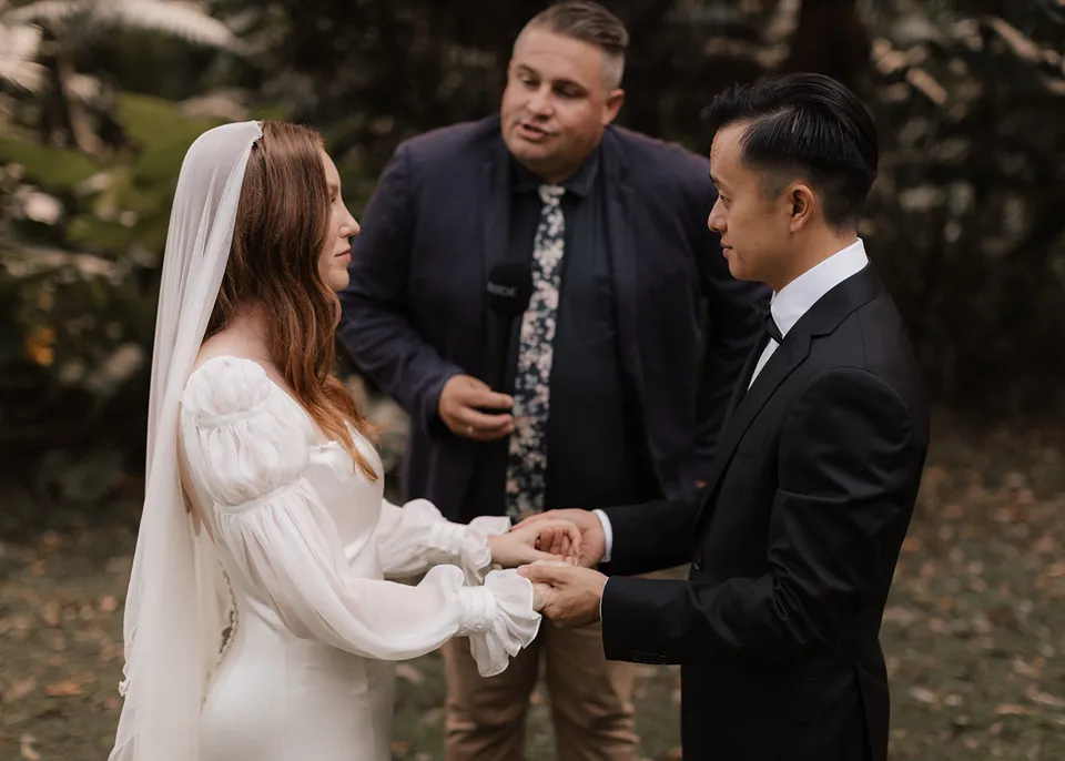 The celebrant speaking with Elyse and Jonathan as they hold hands. At Fig Tree Villa in Ewingsdale, Byron Bay, for an Elopement Collective elopement. Photograph by Bulb Creative.
