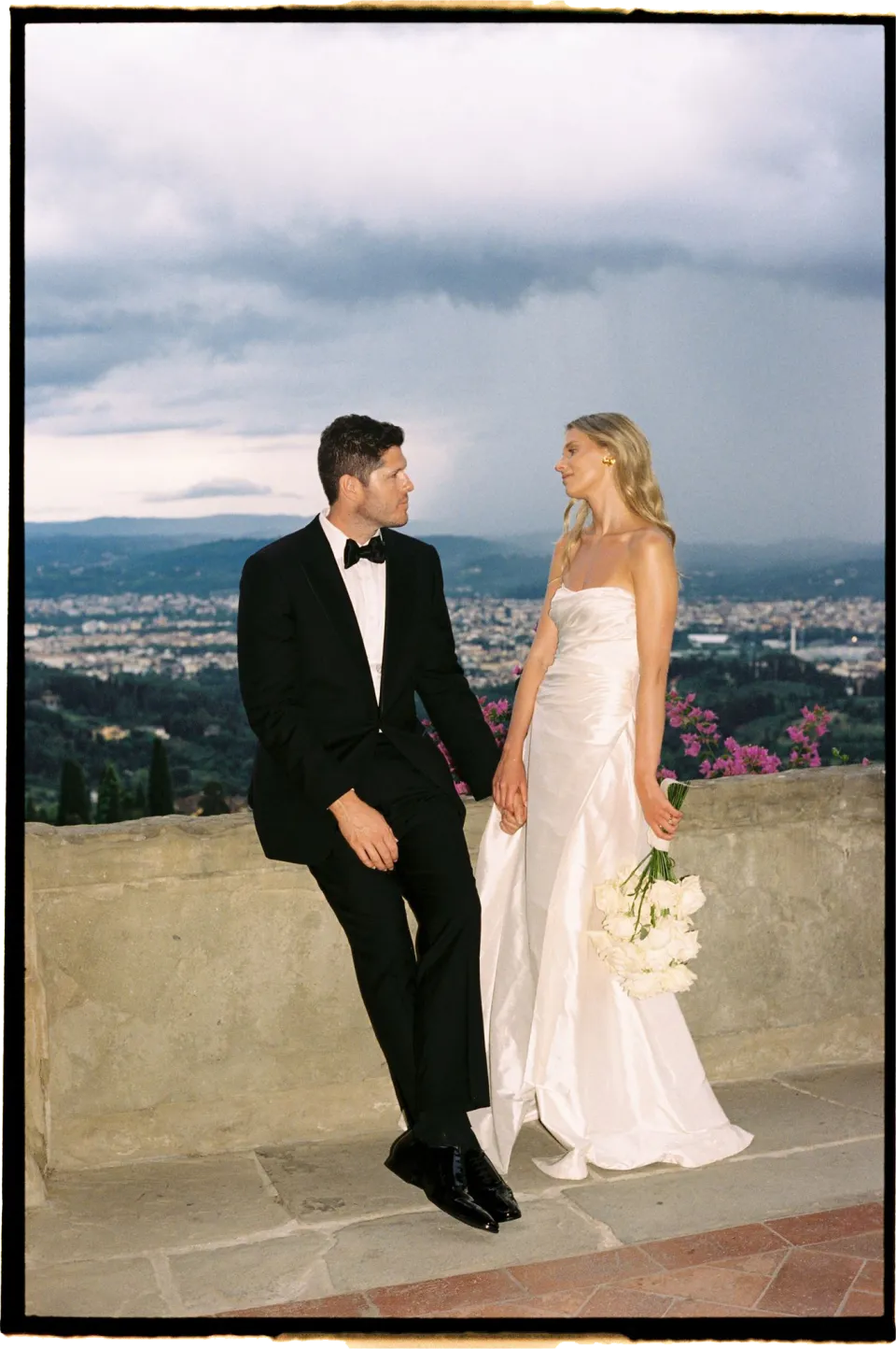 Holly and Matthew smiling at each other beside the terrace balustrade, with her white bouquet by Le Francesche Flower Designers and Florence behind them, at Villa San Michele in Fiesole above Florence, Tuscany, Italy, for an Elopement Collective elopement. Film photograph by Khara from House of Lucie.