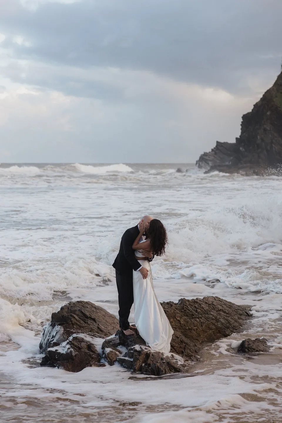 Rachel and Dane embracing on the rocks beneath a cloud-filled coastal sky. At Whites Beach, Broken Head, near Byron Bay, for an Elopement Collective elopement. Photograph by Jason Lucas.