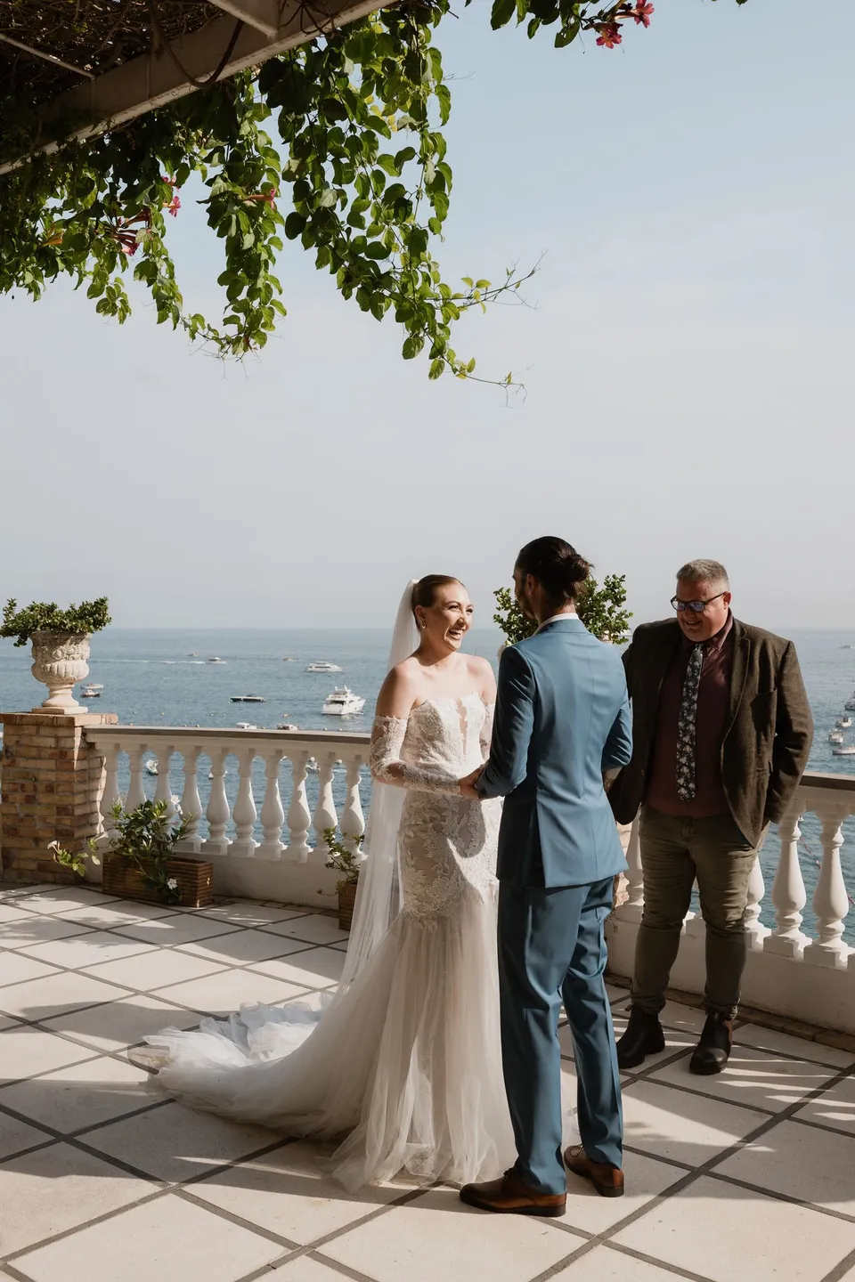 Skyler reading her vows to Zach beside the terrace balustrade, with celebrant Josh Withers standing nearby, at Hotel Buca di Bacco in Positano, Amalfi Coast, Italy, for an Elopement Collective elopement. Photograph by Jason Corroto.