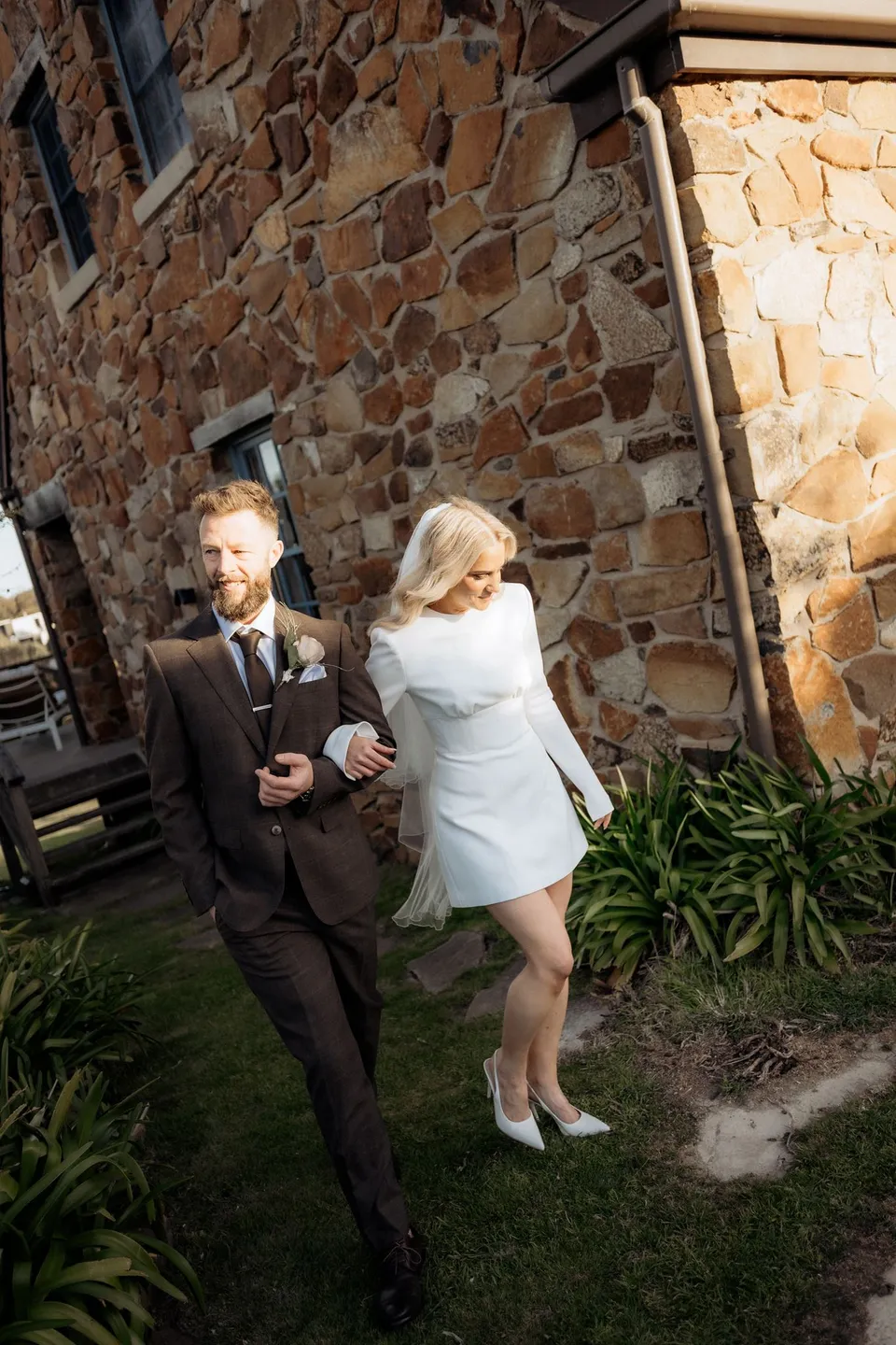 Kaitlyn and Russell walking arm in arm beside a sunlit stone cottage. At Piermont Retreat on Tasmania’s east coast, for an Elopement Collective elopement. Photograph by Omar of By The Wilde.