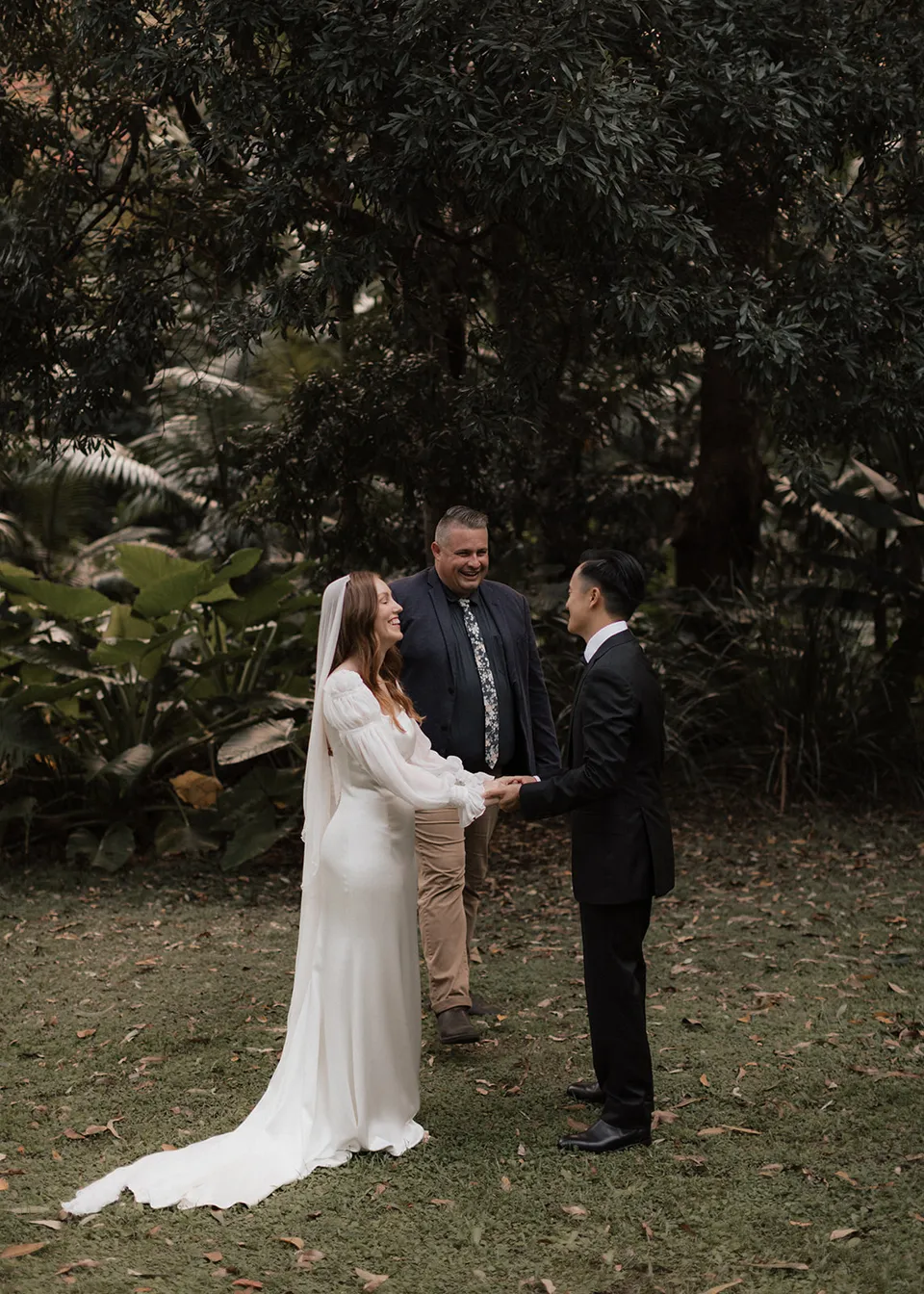 Elyse and Jonathan listening to celebrant Josh Withers beneath the garden trees. At Fig Tree Villa in Ewingsdale, Byron Bay, for an Elopement Collective elopement. Photograph by Bulb Creative.