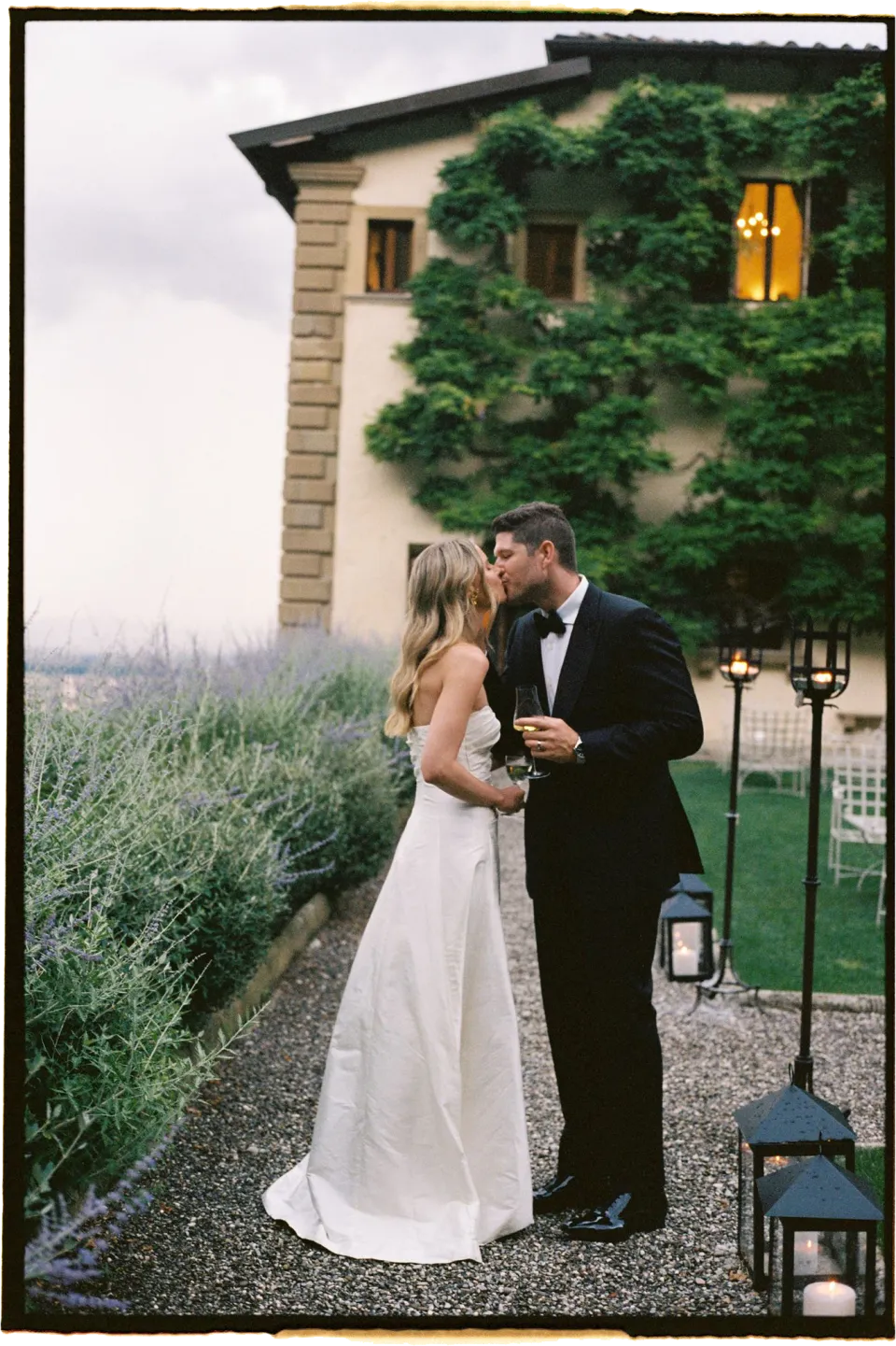 Holly and Matthew kissing beside ivy-covered stone walls and a glowing lantern outside the villa, with her white bouquet by Le Francesche Flower Designers, at Villa San Michele in Fiesole above Florence, Tuscany, Italy, for an Elopement Collective elopement. Film photograph by Khara from House of Lucie.