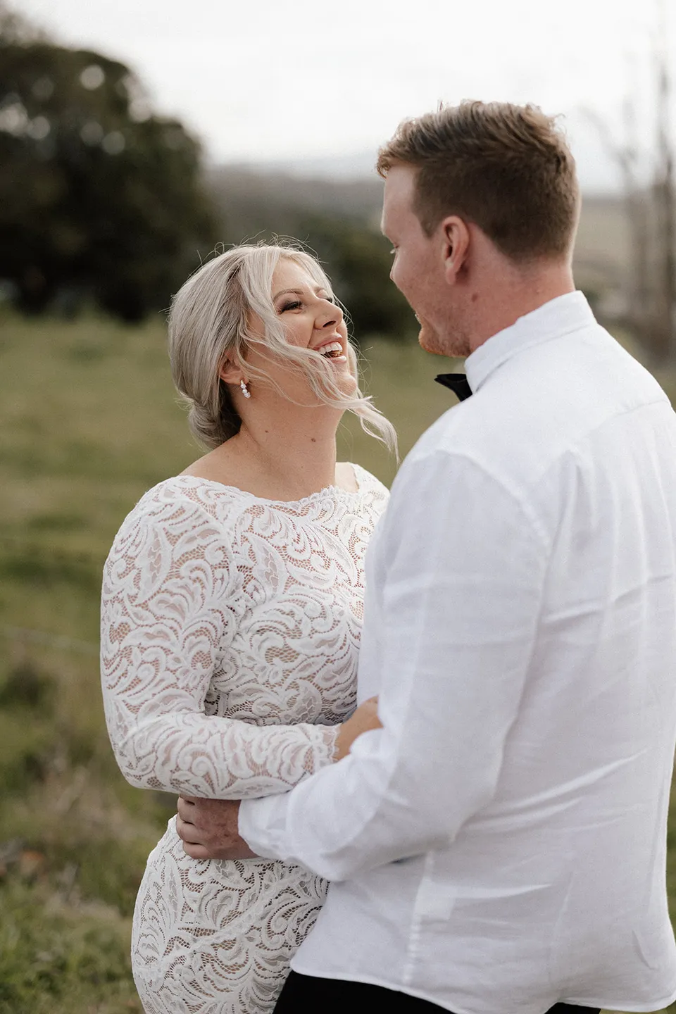 Laura laughing as she faces Matt, with green paddocks behind them, in the Tweed Valley, NSW, for an Elopement Collective elopement. Photo by Bulb Creative.