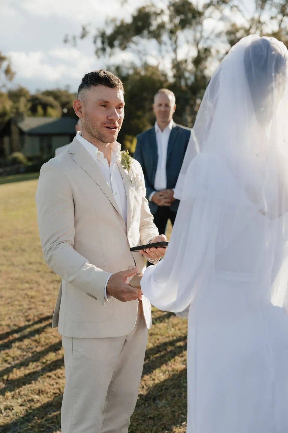 Mitch reading from his phone while holding Renee’s hand during their ceremony, wearing a floral buttonhole by The Romantics. At Piermont Retreat on Tasmania’s east coast, for an Elopement Collective elopement. Photograph by Bulb Creative.