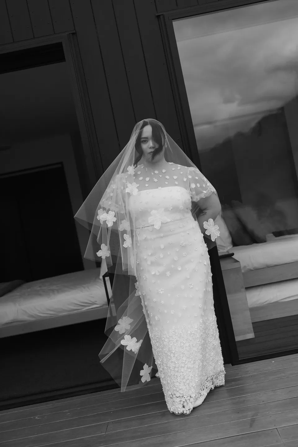 A black and white portrait of Jaeda on the deck in her white dress, sheer daisy-appliqué top and veil, at their alpine Airbnb in Ben Lomond, Queenstown, New Zealand, for an Elopement Collective Queenstown elopement. Photograph by Bulb Creative.