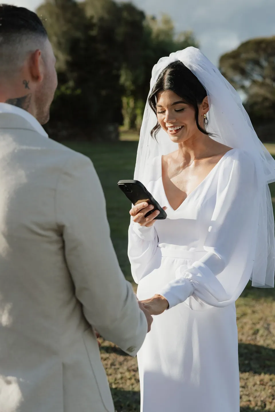 Renee smiling as she reads from her phone to Mitch during their outdoor ceremony. At Piermont Retreat on Tasmania’s east coast, for an Elopement Collective elopement. Photograph by Bulb Creative.