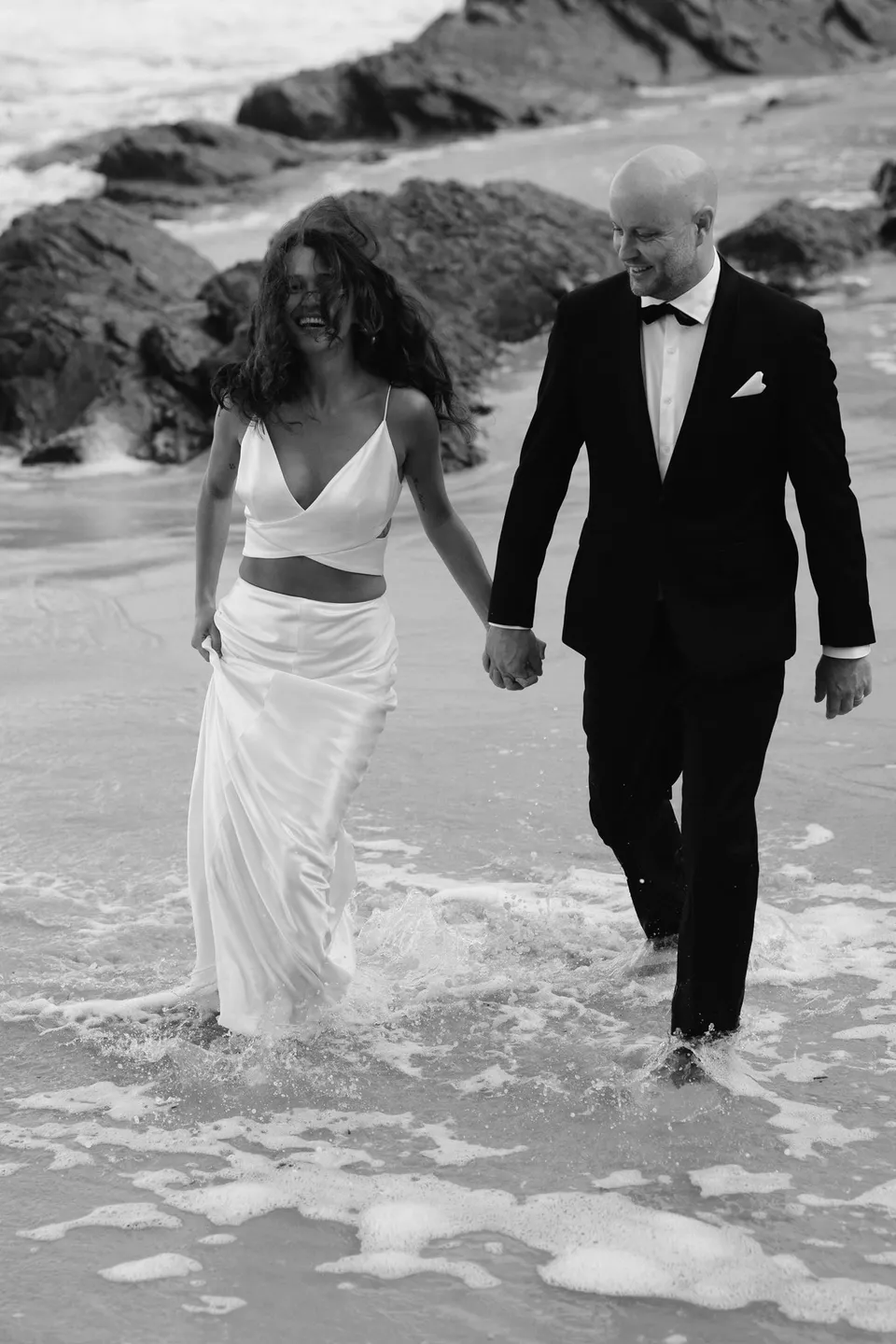 Black-and-white photograph of Rachel and Dane holding hands while wading through the surf. At Whites Beach, Broken Head, near Byron Bay, for an Elopement Collective elopement. Photograph by Jason Lucas.