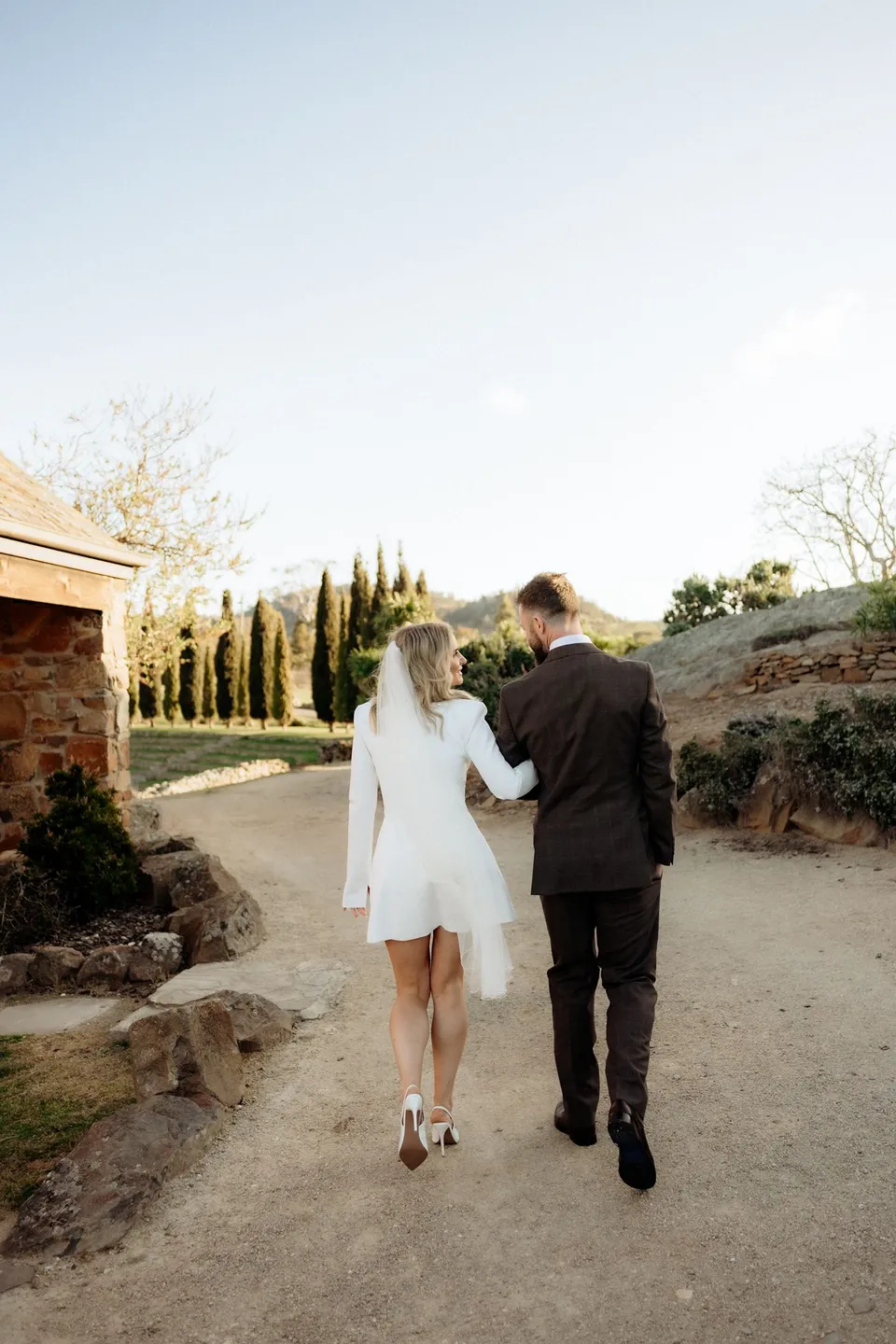 Rear view of Kaitlyn and Russell walking hand in hand towards the cypress trees and stone amphitheatre. At Piermont Retreat on Tasmania’s east coast, for an Elopement Collective elopement. Photograph by Omar of By The Wilde.