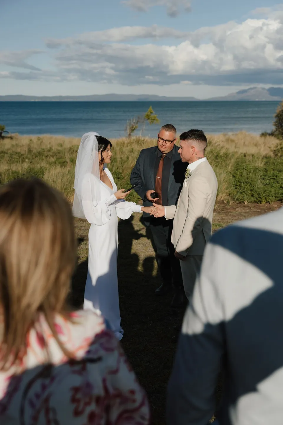 Celebrant Josh Withers leading Renee and Mitch’s ceremony, with their guests gathered close and the bay behind them. At Piermont Retreat on Tasmania’s east coast, for an Elopement Collective elopement. Photograph by Bulb Creative.