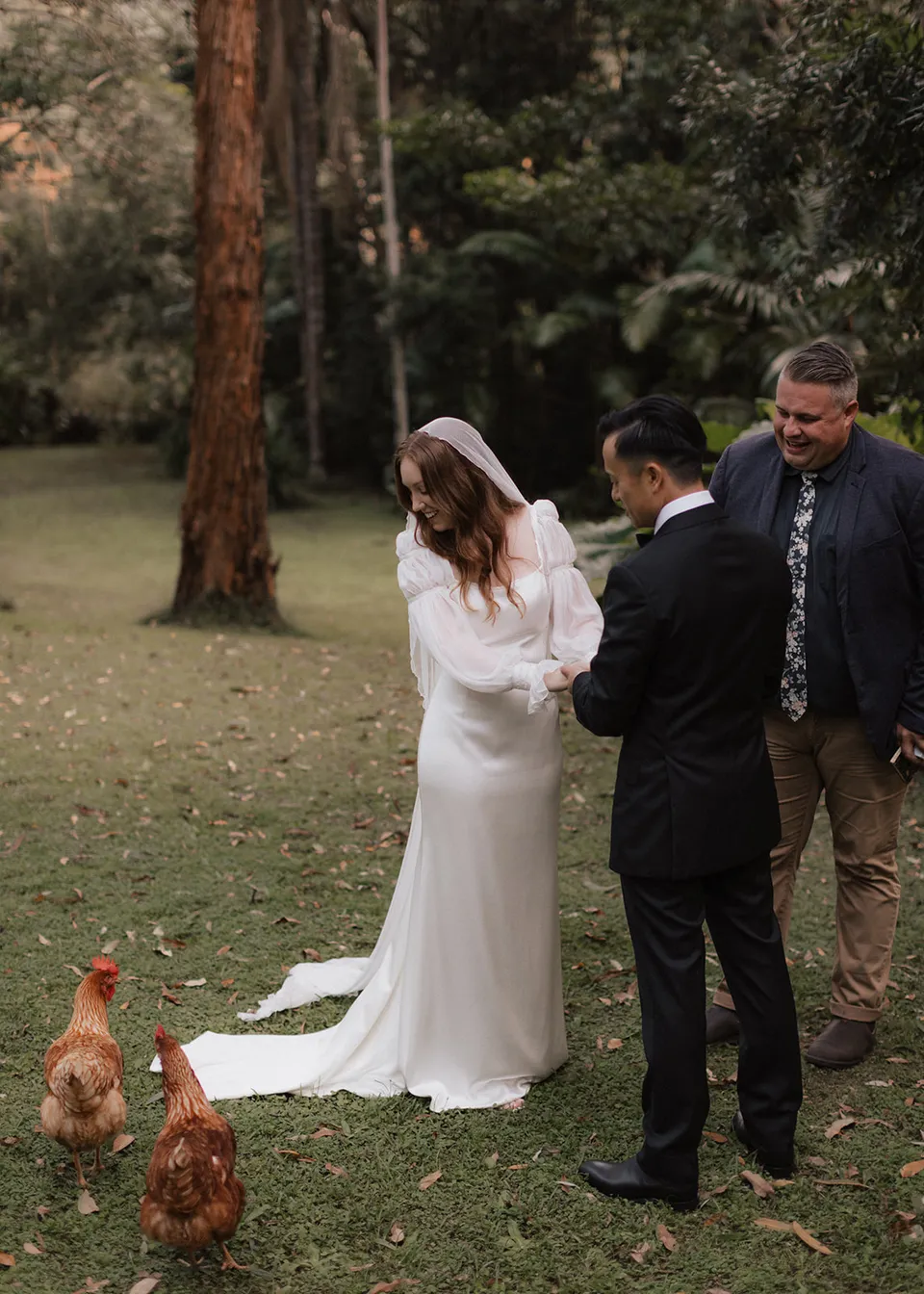 The couple and celebrant Josh Withers looking towards hens beside Elyse's dress. At Fig Tree Villa in Ewingsdale, Byron Bay, for an Elopement Collective elopement. Photograph by Bulb Creative.