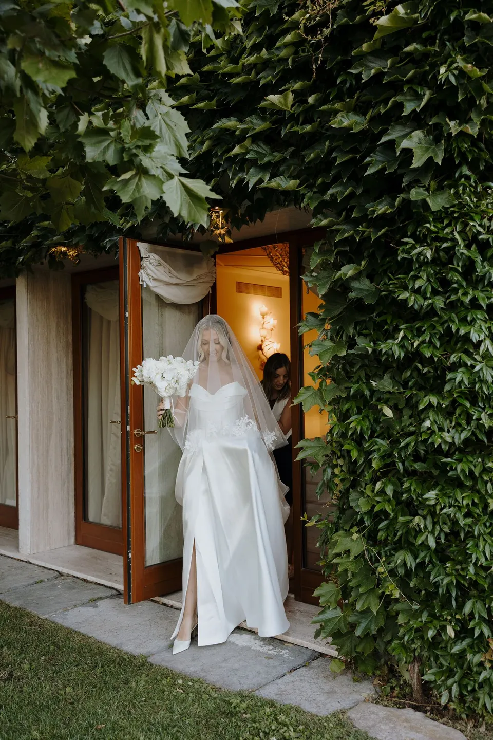 Zoe holding her white bouquet by Garden of Love in an open doorway framed by climbing greenery, at Passalacqua in Moltrasio, Lake Como, Italy, for an Elopement Collective elopement. Photo by George Bowden.