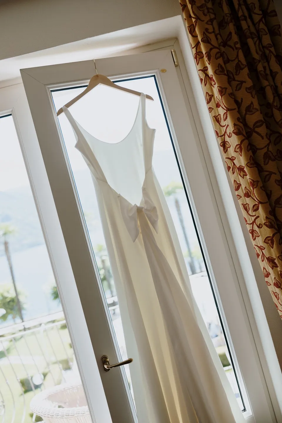 Lyndal’s white wedding dress hanging from a wooden hanger beside a balcony window at Hotel Villa Marie in Tremezzo, at Lake Como in Lombardy, Italy, for an Elopement Collective elopement. Photograph by George Bowden.