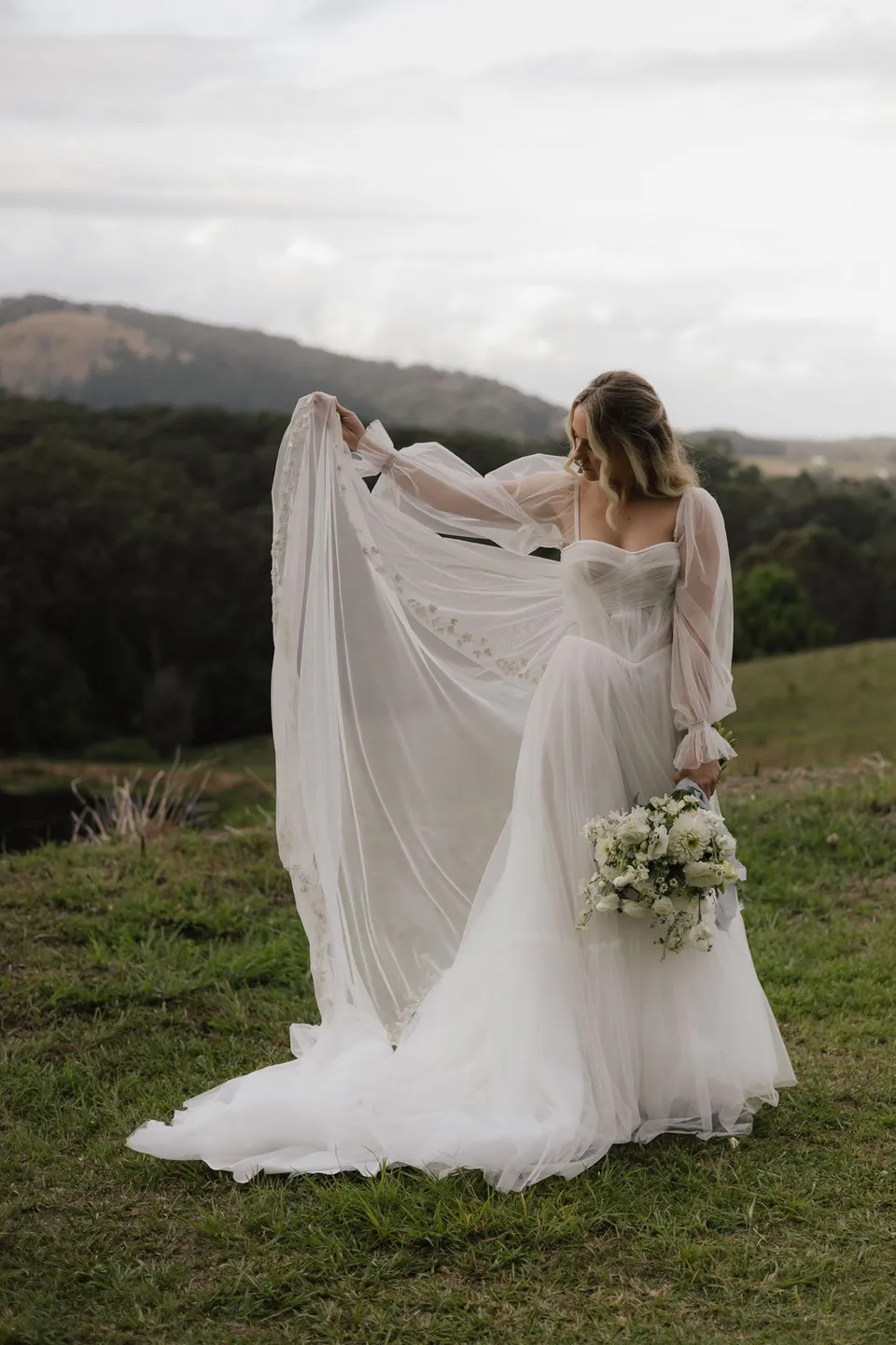 Bridget lifting her veil against a backdrop of green hills. At Cudgera Creek in northern New South Wales, for an Elopement Collective elopement. Photograph by Bulb Creative.