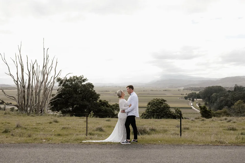 Matt and Laura standing together beside a country road overlooking rolling hills and paddocks, in the Tweed Valley, NSW, for an Elopement Collective elopement. Photo by Bulb Creative.
