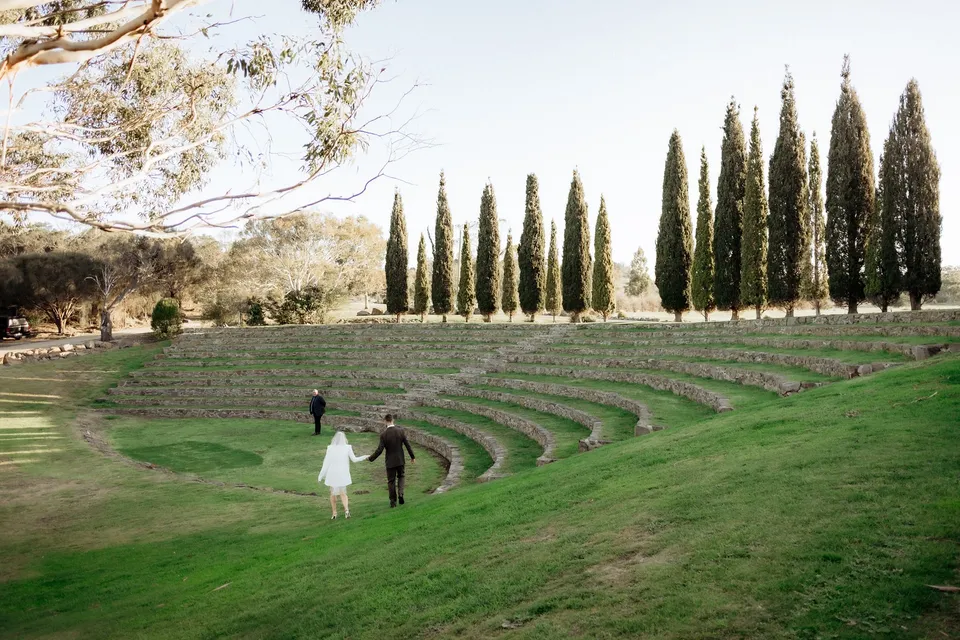 Wide view of Kaitlyn and Russell walking into the grass and stone amphitheatre for their ceremony. At Piermont Retreat on Tasmania’s east coast, for an Elopement Collective elopement. Photograph by Omar of By The Wilde.