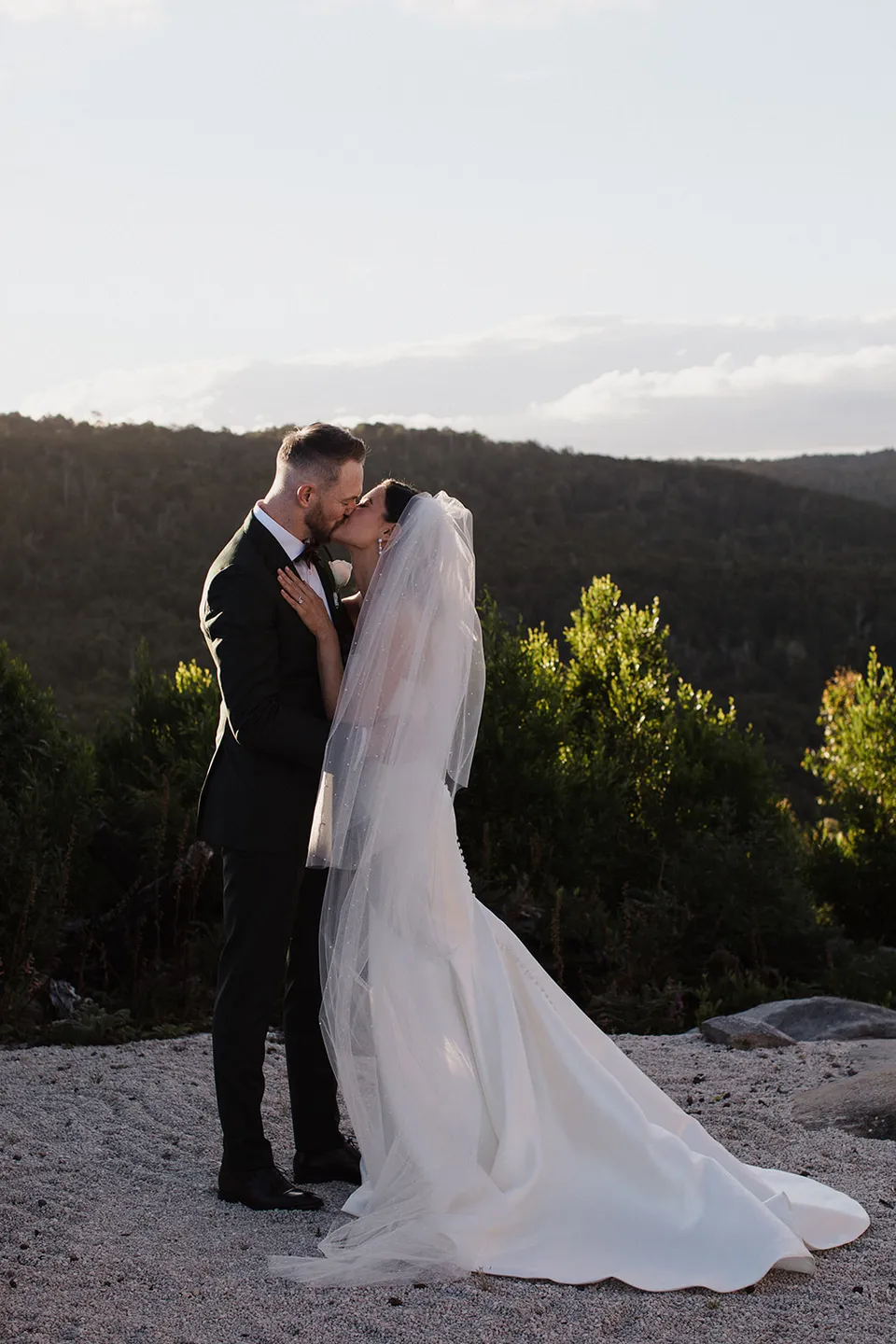 Meg and Ryan kissing after their ceremony, with her train spread across the rock and the forest behind them. At The Keep on Tasmania’s east coast, for an Elopement Collective elopement. Photograph by Jenna Agius.