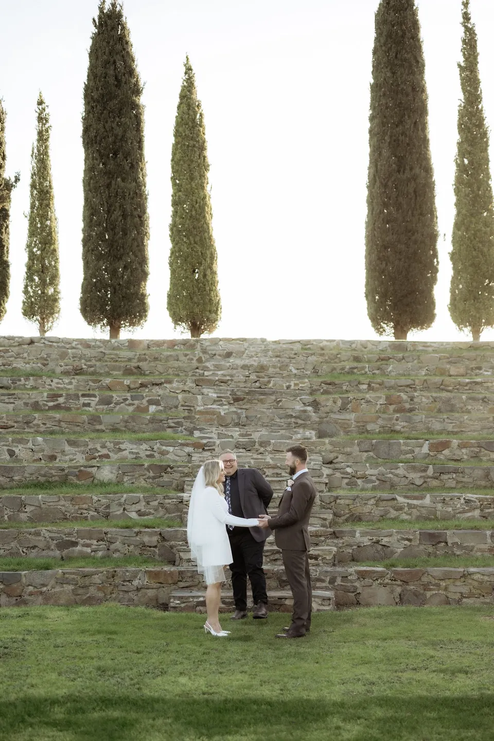 Kaitlyn and Russell holding hands as Josh Withers leads their ceremony beneath the cypress trees. At Piermont Retreat on Tasmania’s east coast, for an Elopement Collective elopement. Photograph by Omar of By The Wilde.
