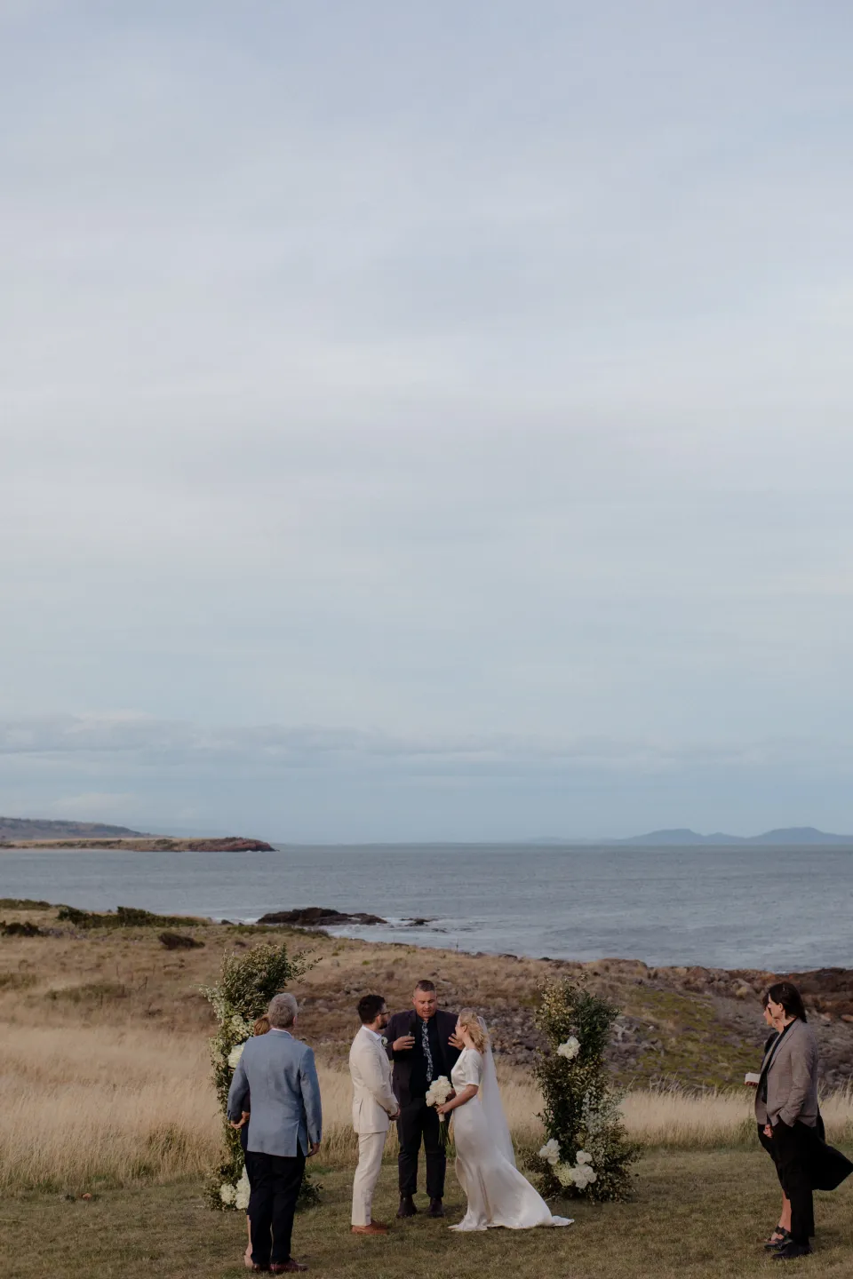 Jemmah and Jack’s ceremony beneath a broad sky with the coast beyond, at Thalia Haven on the East Coast of Tasmania, Australia, for an Elopement Collective elopement. Photograph by Joey Willis.