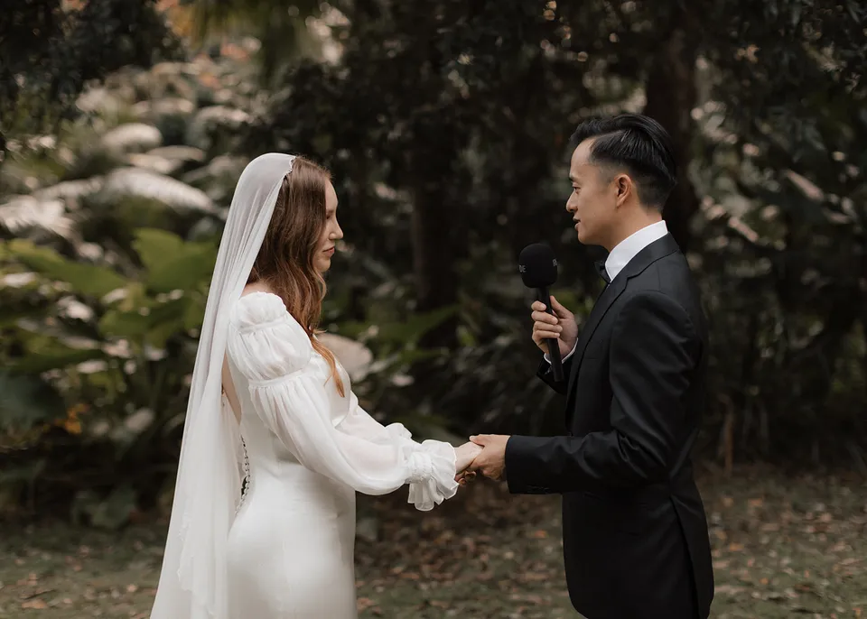 Jonathan holding a microphone and saying his vows to Elyse as they hold hands beneath the garden canopy at Fig Tree Villa in Ewingsdale, Byron Bay. An Elopement Collective elopement, photographed by Bulb Creative.