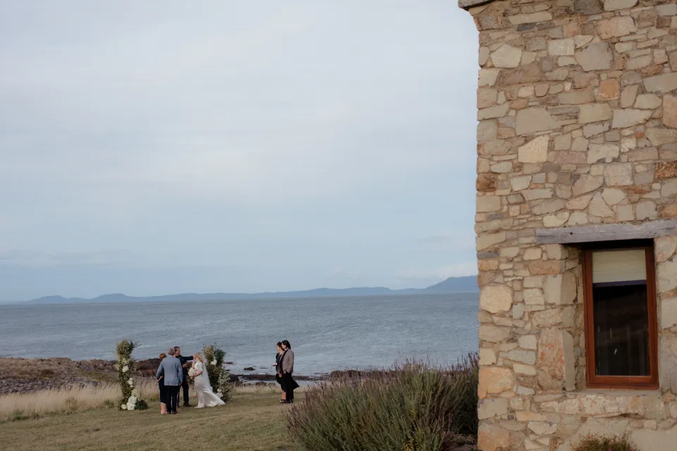 The stone house framing the small ceremony on the coastal lawn, at Thalia Haven on the East Coast of Tasmania, Australia, for an Elopement Collective elopement. Photograph by Joey Willis.