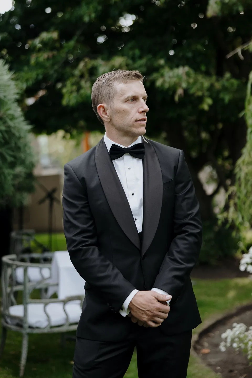 Andrew in a black tuxedo and bow tie, standing beneath leafy trees beside the ceremony chairs, at Islington Hotel in South Hobart, Tasmania, Australia, for an Elopement Collective summer elopement. Photograph by George Bowden.