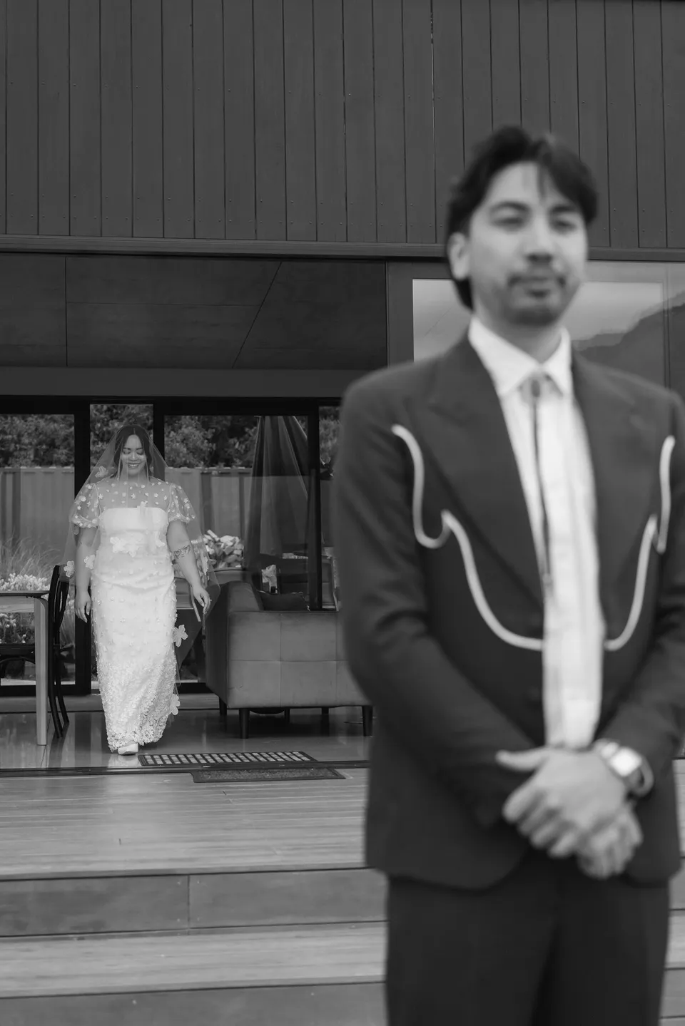 A black and white photograph of Adrian waiting outside while Jaeda approaches from the open doorway for their first look, at their alpine Airbnb in Ben Lomond, Queenstown, New Zealand, for an Elopement Collective Queenstown elopement. Photograph by Bulb Creative.