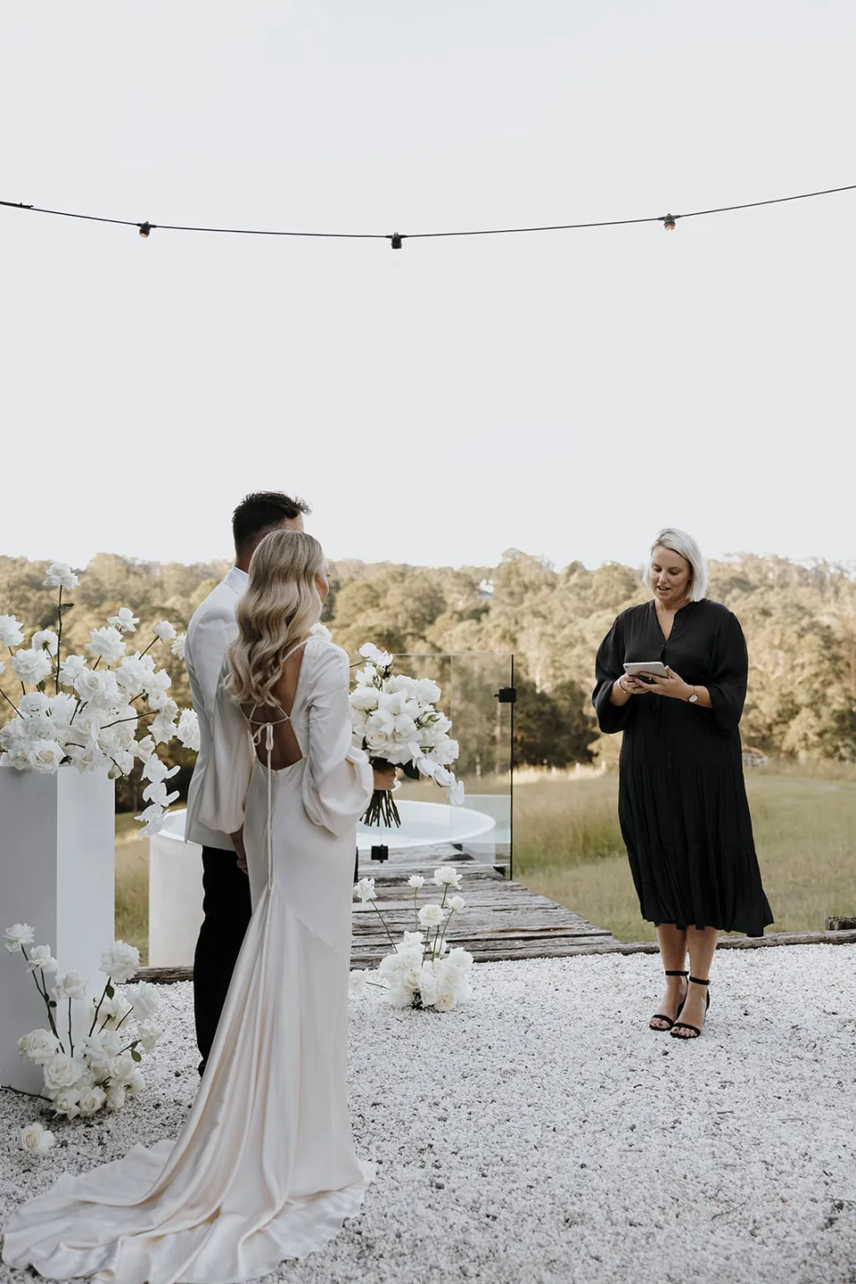 Hayley Hart leading Gabrielle and Greg’s outdoor ceremony between white floral plinths beneath festoon lights at Roundhill Retreat in Obi Obi, Sunshine Coast Hinterland, for their Elopement Collective elopement.