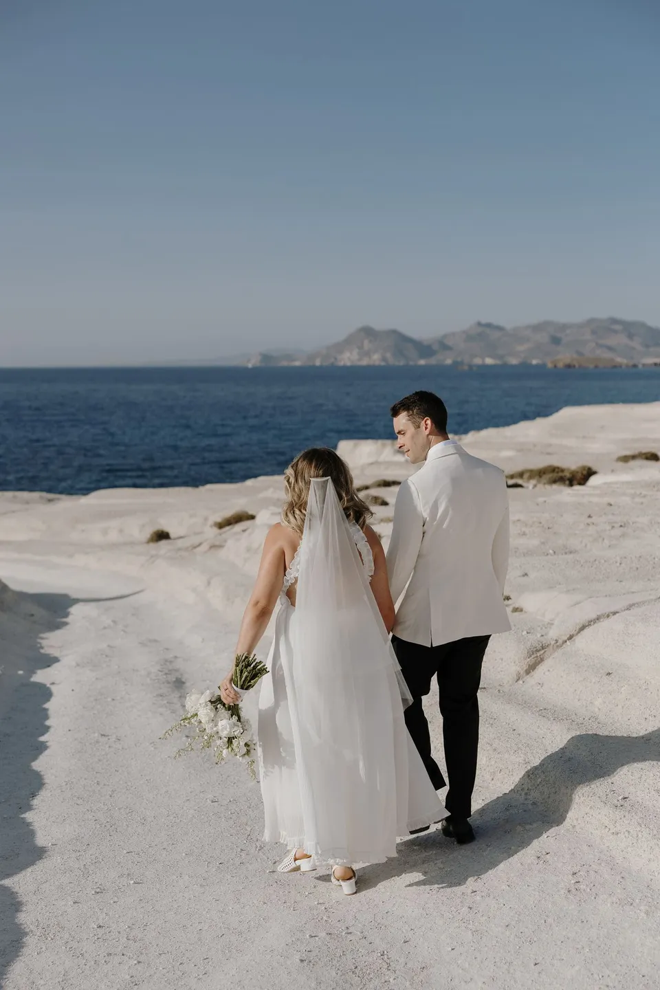 Georgina and Brad holding hands and walking towards the sea across white rocks, with her Giasemi bouquet, at Sarakiniko on Milos in the Cyclades, Greece, during an Elopement Collective elopement. Photograph by George Bowden.