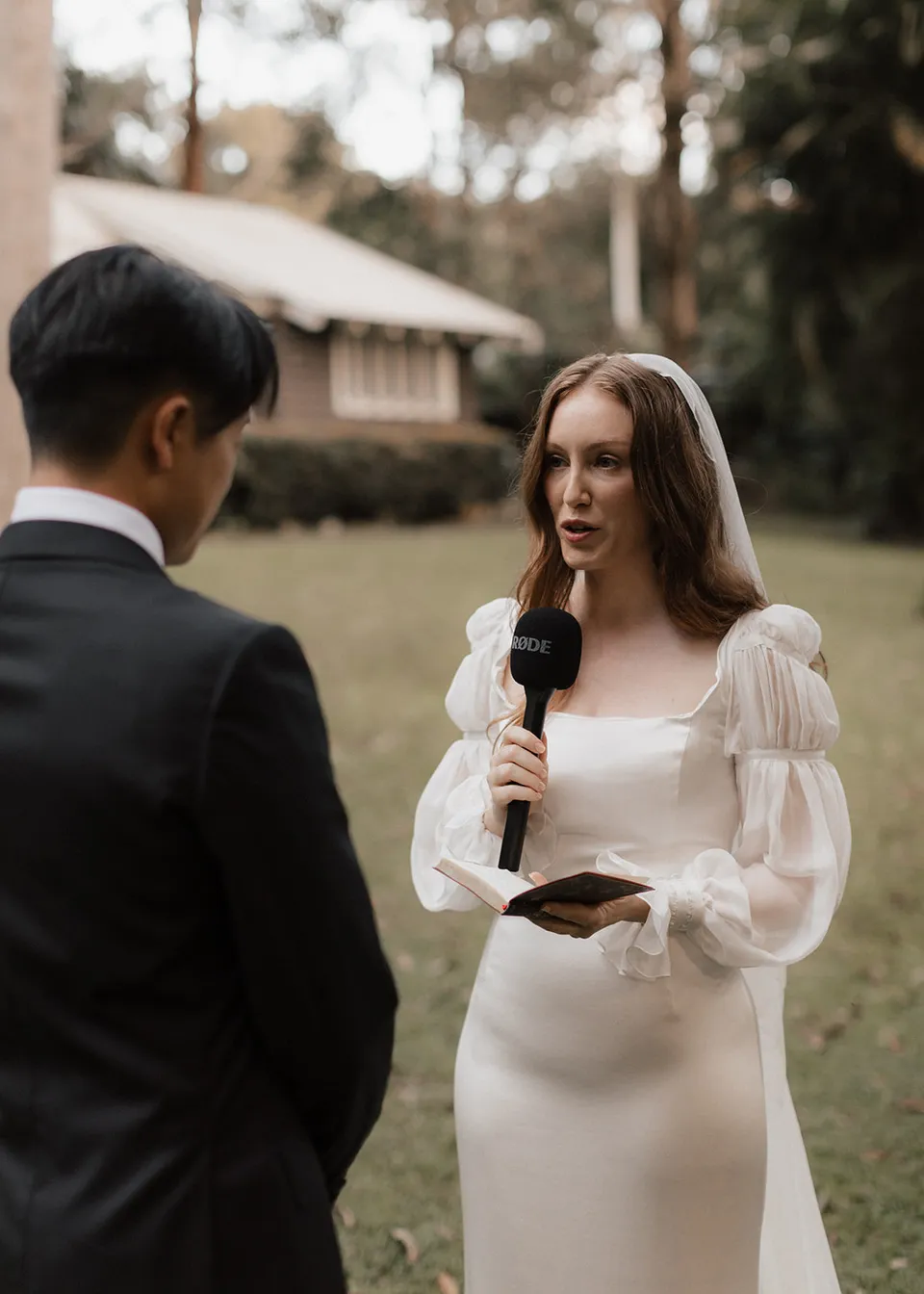 Elyse reading her vows from a small booklet while holding the microphone. At Fig Tree Villa in Ewingsdale, Byron Bay, for an Elopement Collective elopement. Photograph by Bulb Creative.