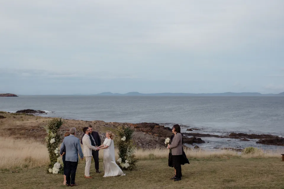 Jemmah and Jack standing with Josh Withers and their loved ones for the ceremony, at Thalia Haven on the East Coast of Tasmania, Australia, for an Elopement Collective elopement. Photograph by Joey Willis.