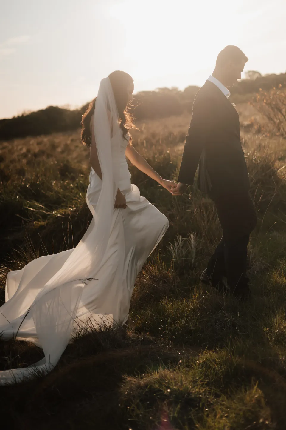 Dani and Anthony walking hand in hand through coastal grass, with warm sunlight catching her veil and dress, at Thalia Haven on Tasmania’s east coast, for an Elopement Collective elopement. Photograph by Bulb Creative.
