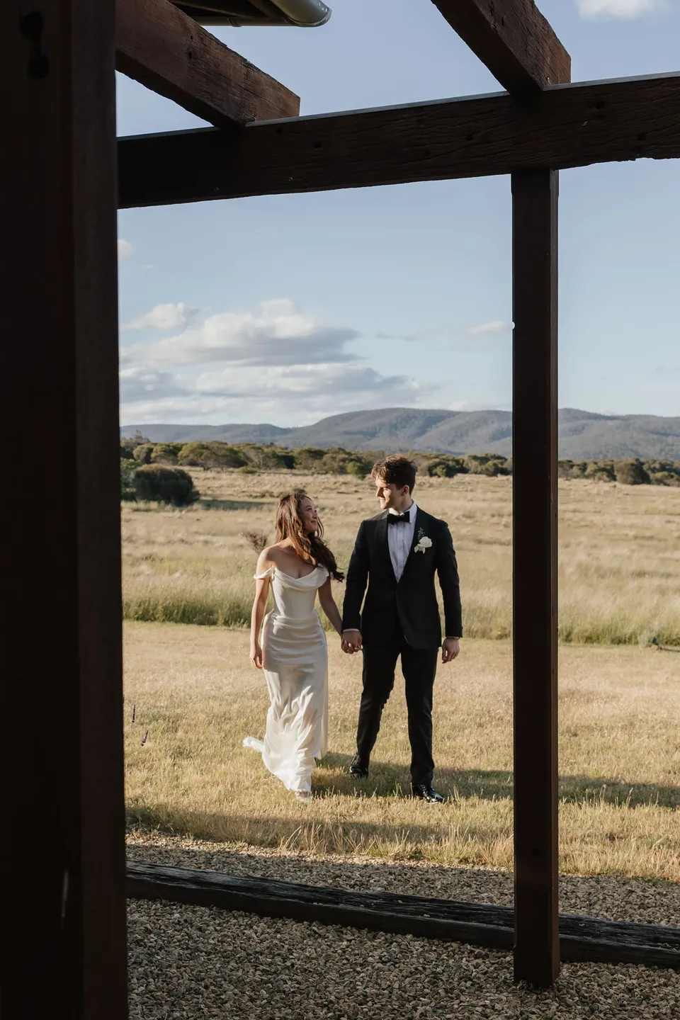 Clara and Harry walking hand in hand towards a timber pergola, with coastal grassland behind them, at Thalia Haven on the East Coast of Tasmania, for an Elopement Collective elopement. Photograph by Bulb Creative.