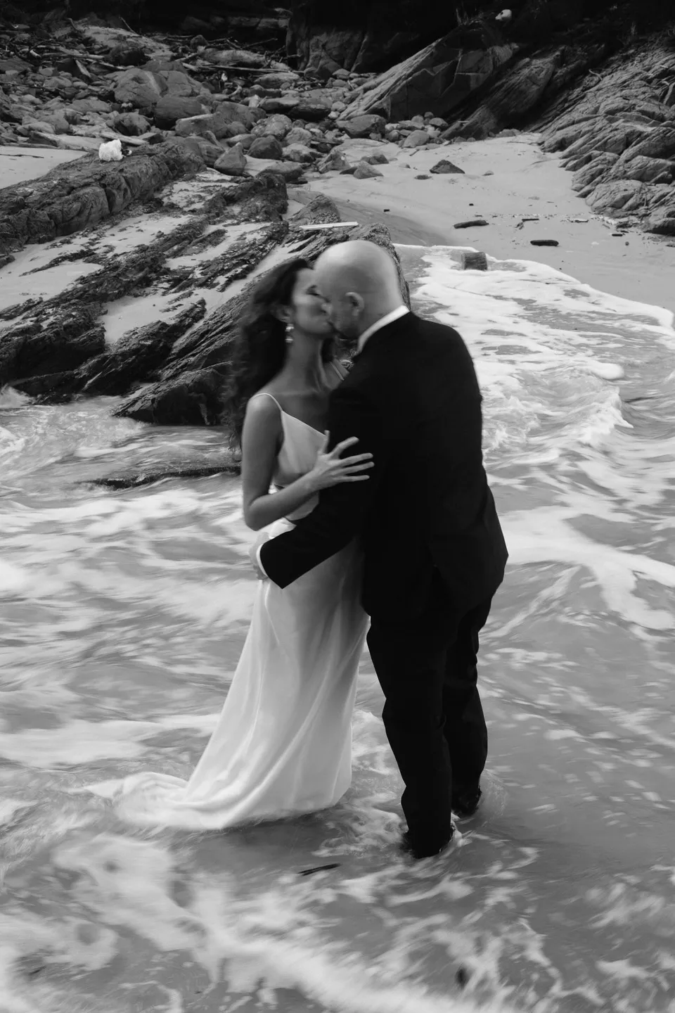 Black-and-white photograph of Rachel and Dane kissing as white water washes around them. At Whites Beach, Broken Head, near Byron Bay, for an Elopement Collective elopement. Photograph by Jason Lucas.
