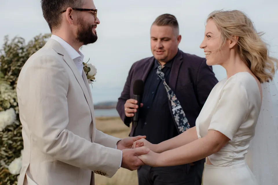 Jemmah and Jack holding hands while Josh Withers leads their ceremony, at Thalia Haven on the East Coast of Tasmania, Australia, for an Elopement Collective elopement. Photograph by Joey Willis.