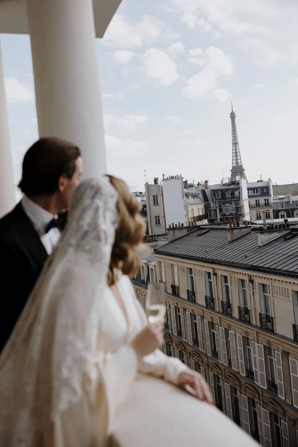 Kara and Samuel looking across Parisian rooftops towards the Eiffel Tower from the hotel balcony, at Hotel Marignan Champs-Élysées in Paris, France, during an Elopement Collective elopement. Photograph by George Bowden.